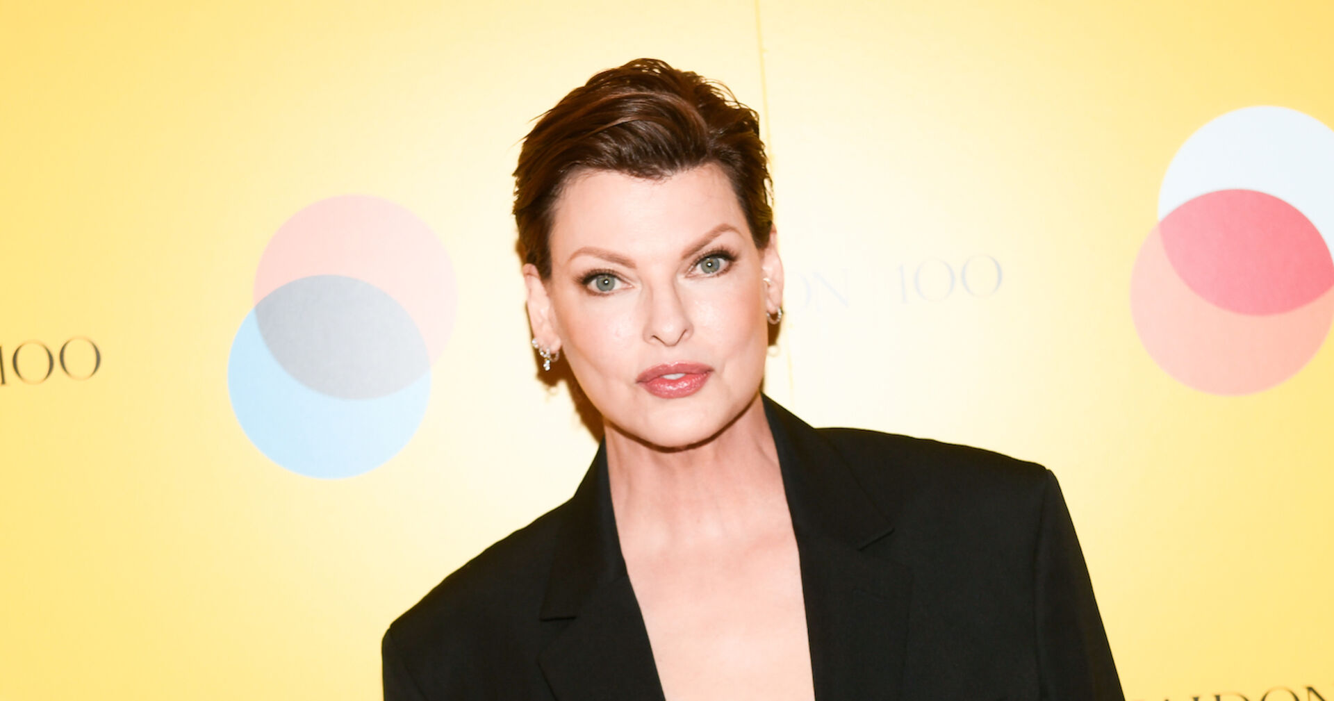 Linda Evangelista mit kurzen Haaren und einem schwarzen Anzug vor einer gelben Wand