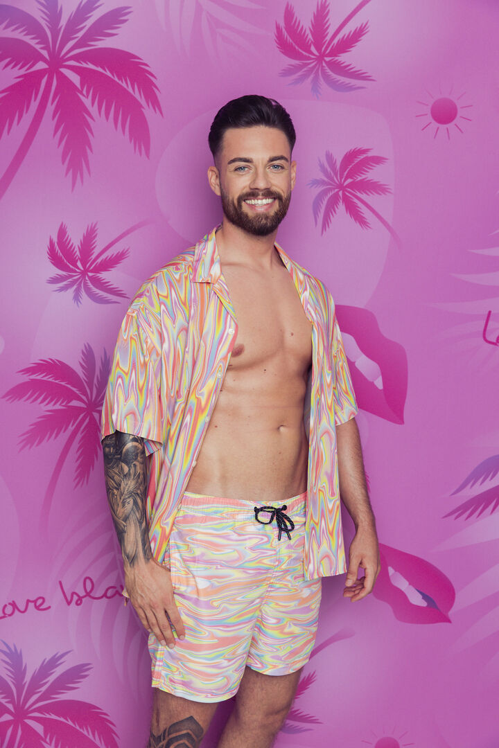 Mann mit bunten Hemd und dazu passender bunter Hose und kurzen braunen Haaren vor einer pinken Wand