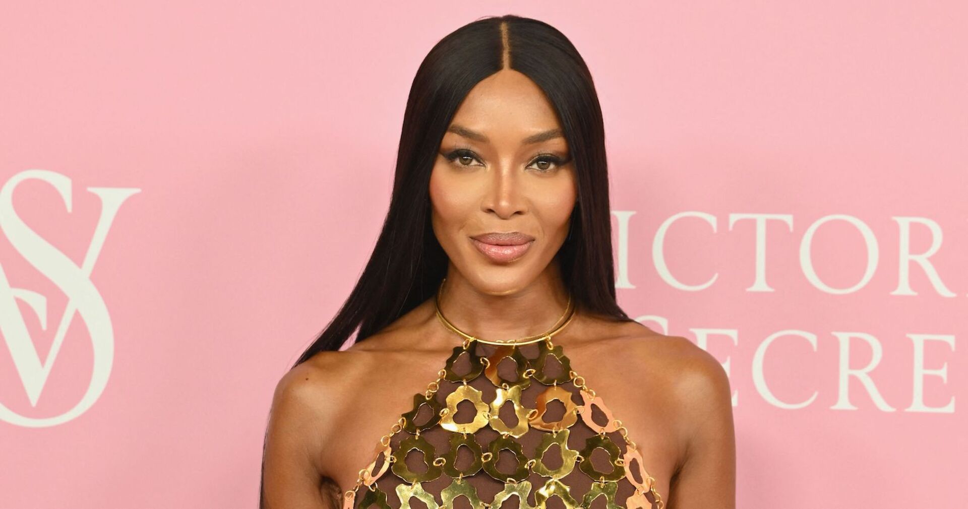 Naomi Campbell mit langen schwarzen Haaren und einem goldenen Kleid vor einer rosa Victoria Secrets Wand.