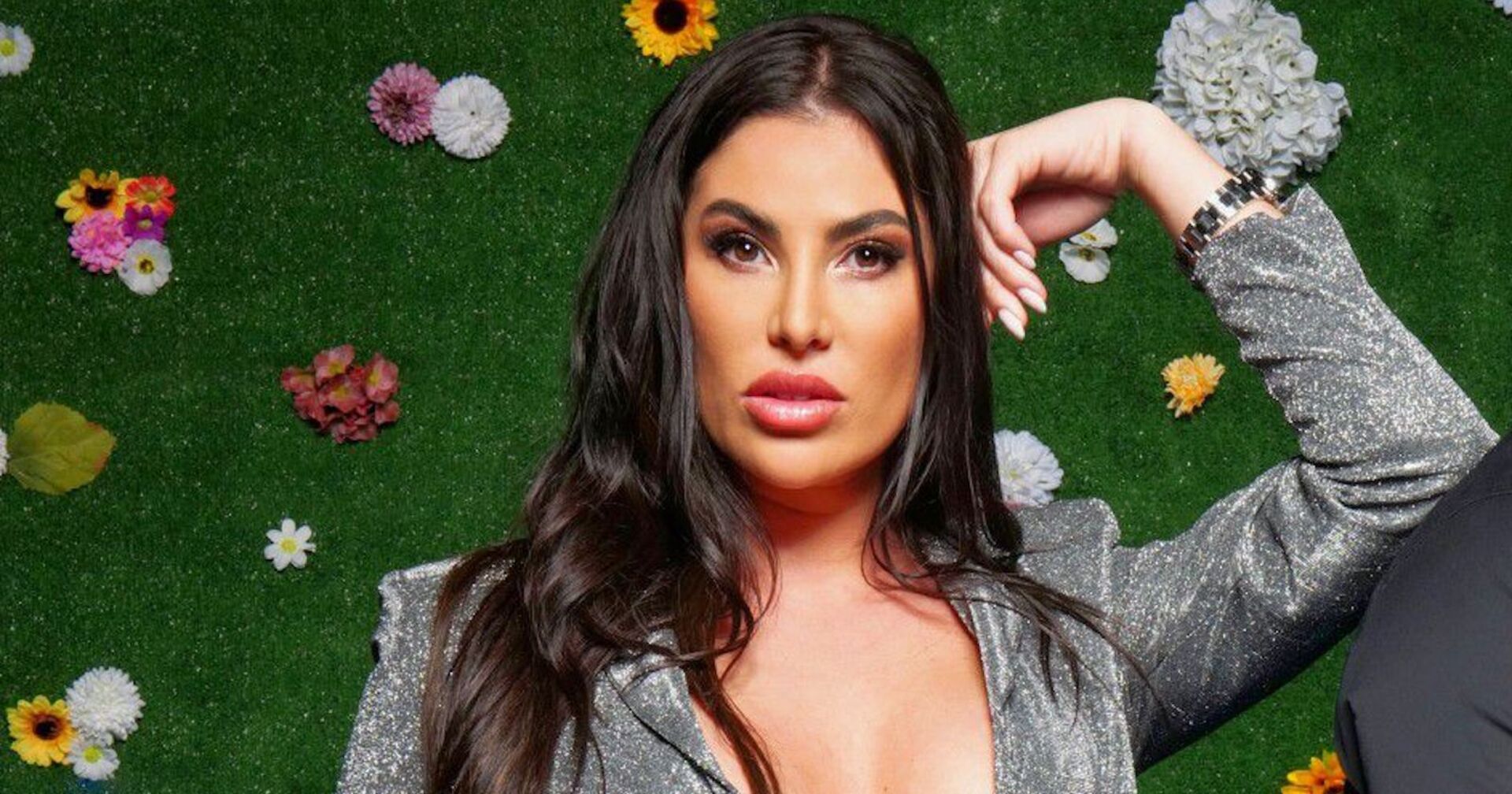 &quot;Sommerhaus der Stars&quot;-Teilnehmerin Ricarda Raatz in einem silbernen Kleid mit schwarzen Haaren vor einer grünen Wand mit Blumen.