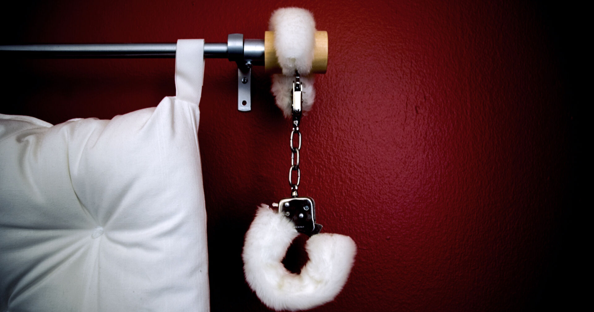 Ein Bett vor einer weinroten Wand. Am Bettgestell hängt eine Handschelle.