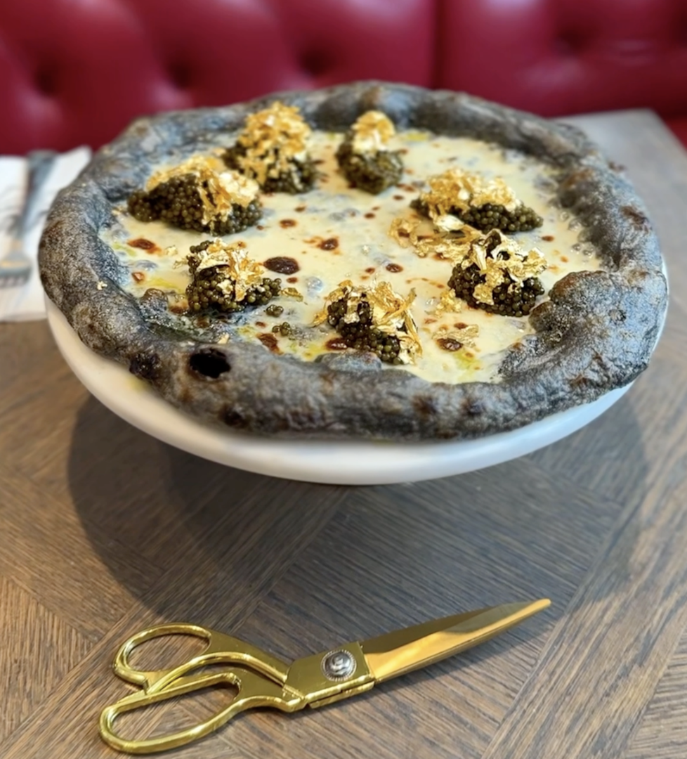 Eine violette Pizza mit Kaviar und Blattgold auf einem weißen Teller. Dabei liegt eine goldene Schere.