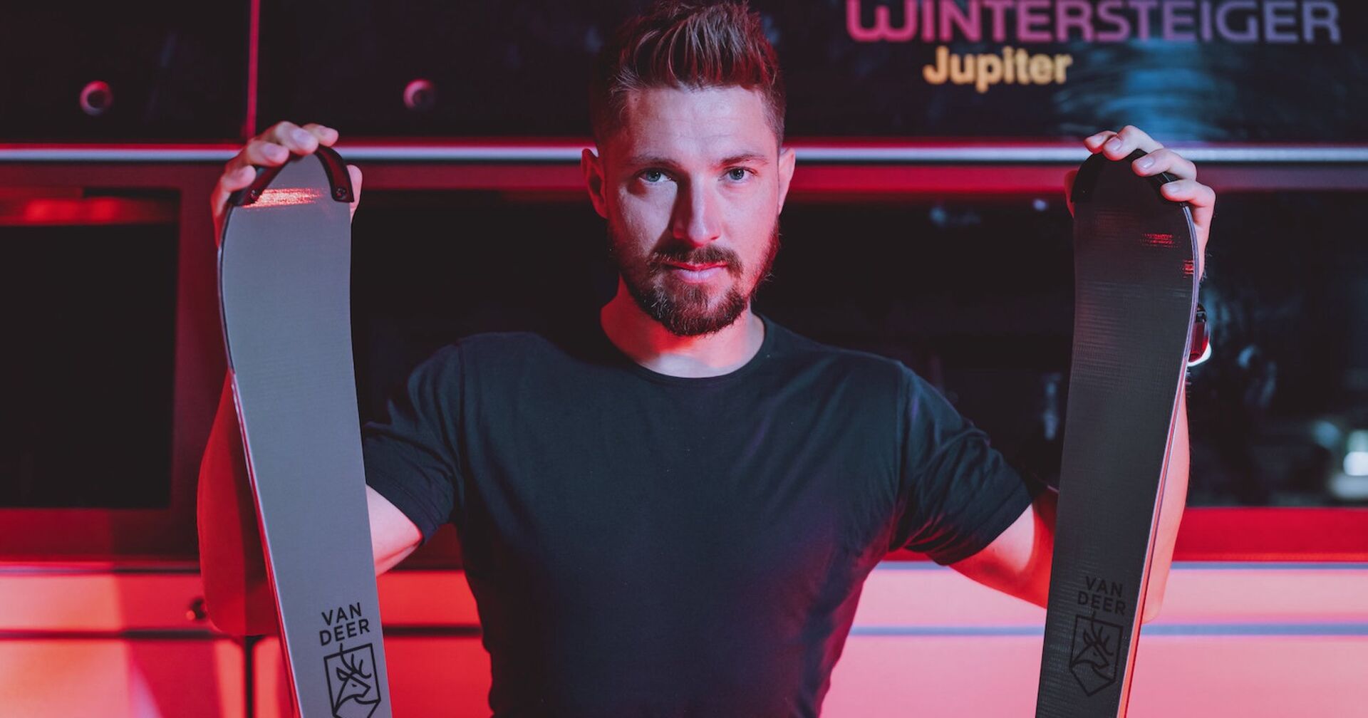 Marcel Hirscher in einem schwarzen T-Shirt, der zwei Prototypen seiner Van-Deer-Skier in die Kamera hält