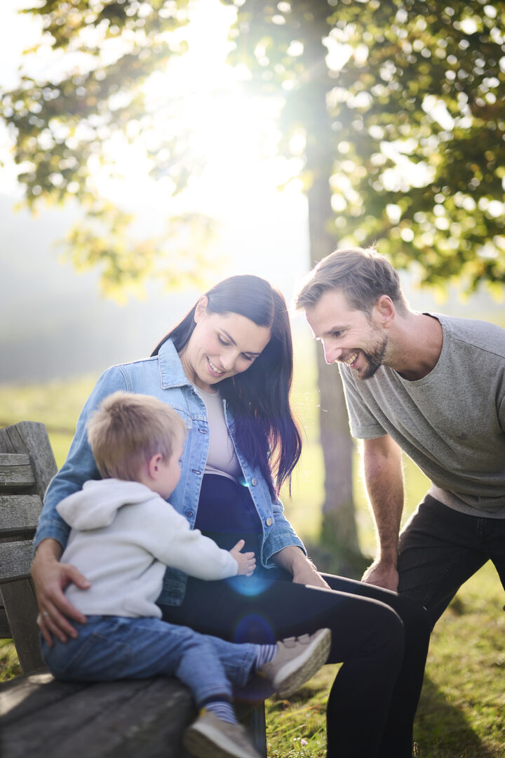 Anna Veith mit offenen schwarzen Haaren und ihr Ehemann Manuel Veith mit kurzen Haaren und einem Bart. Beide schauen auf ihren dreijährigen Sohn Henry, der sanft Annas Schwangerschaftsbauch berührt.