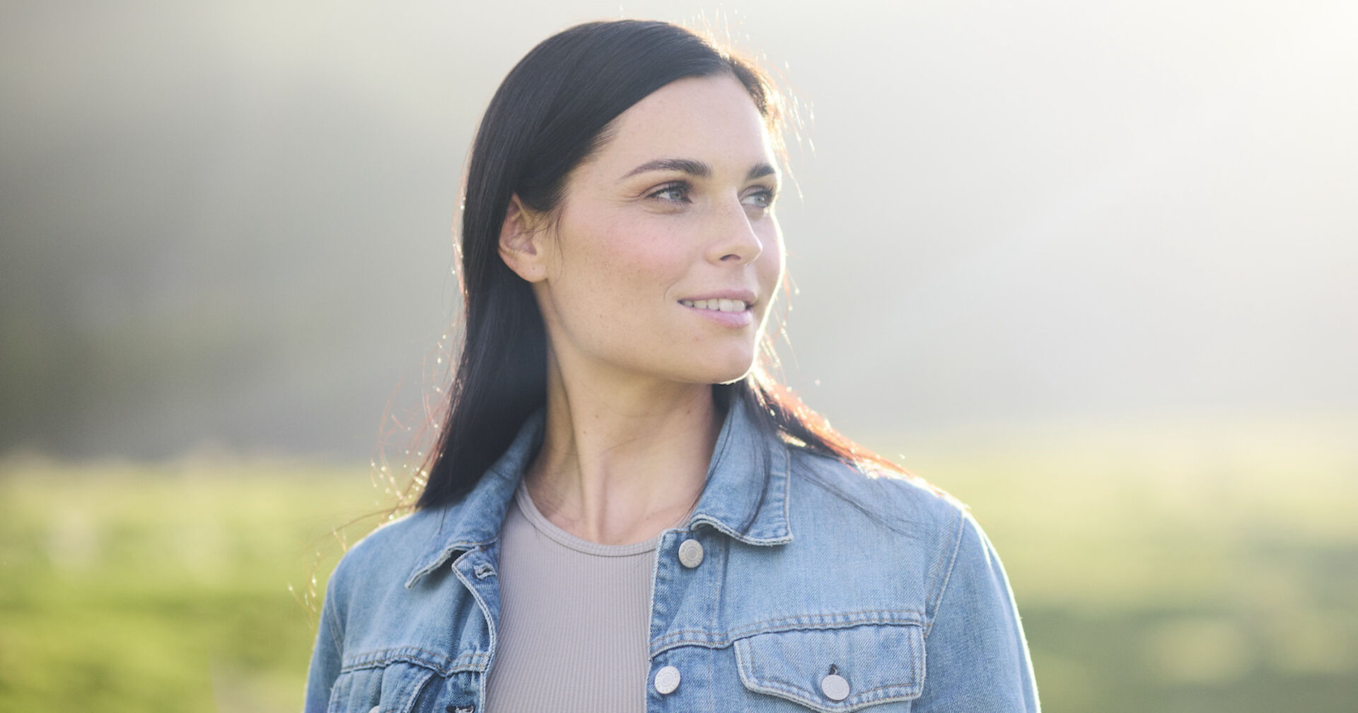 Anna Veith mit offenen schwarzen Haaren und einer blauen Jeansjacke vor einer grünen Wiese.