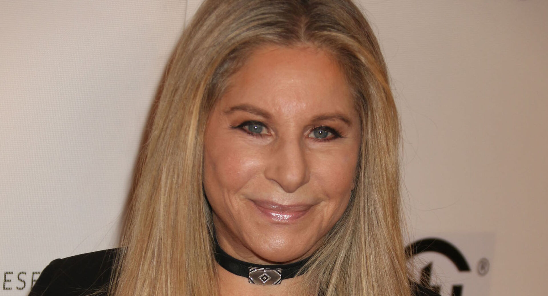 Barbara Streisand mit einer engen schwarzen Kette und offenen blonden Haaren vor einer Fotoleinwand.