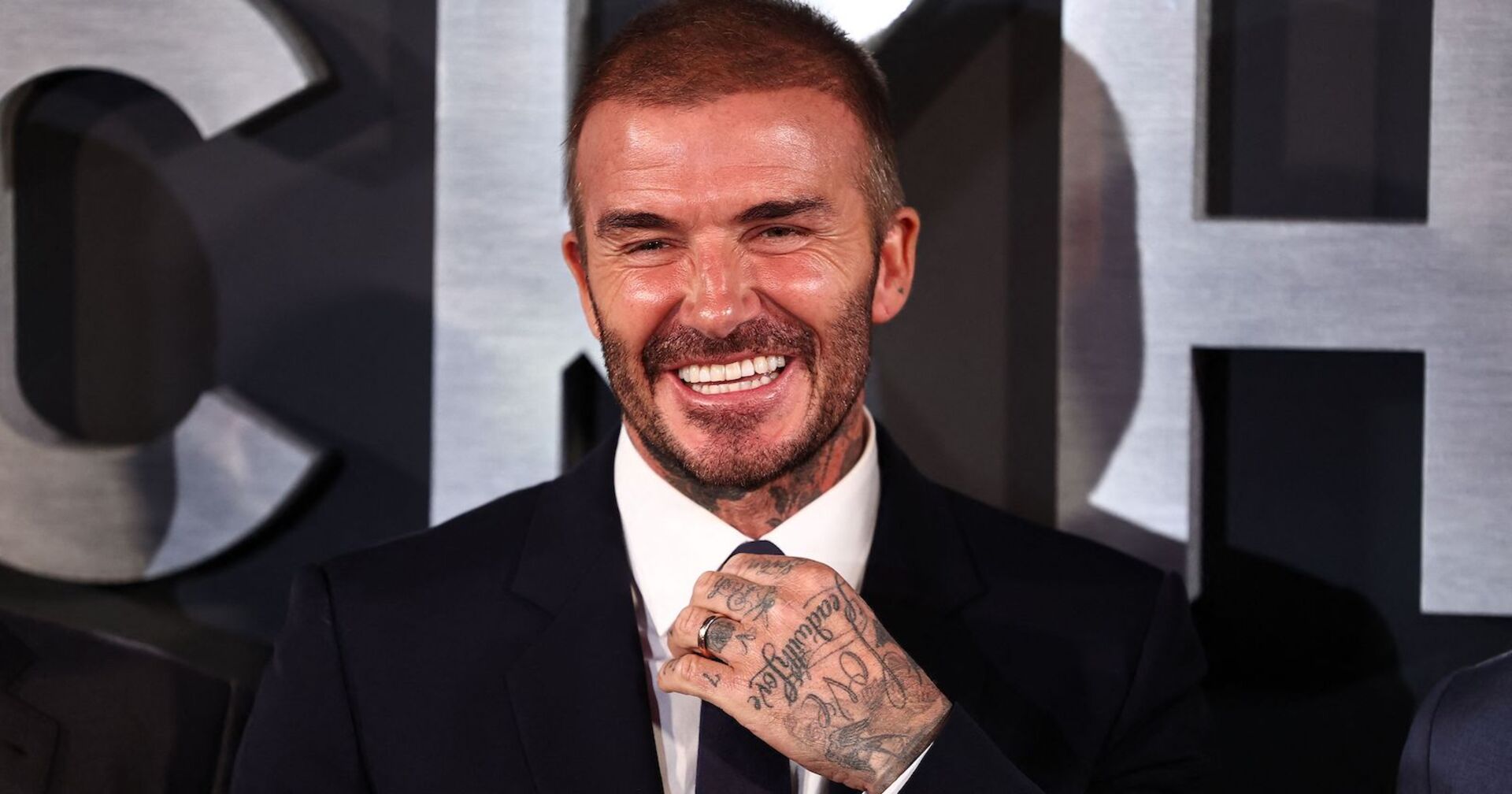 David Beckham lacht in die Kamera und richtet seine Krawatte.