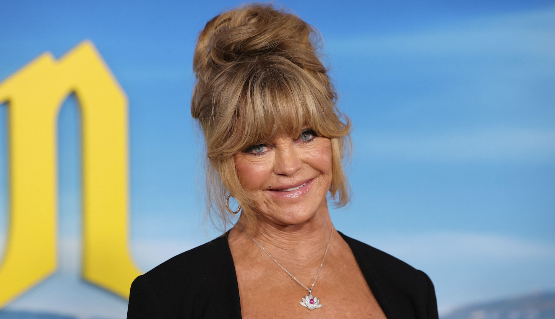 Schauspielerin Goldie Hawn trägt hochgesteckte Haare und steht vor einer blauen Leinwand.