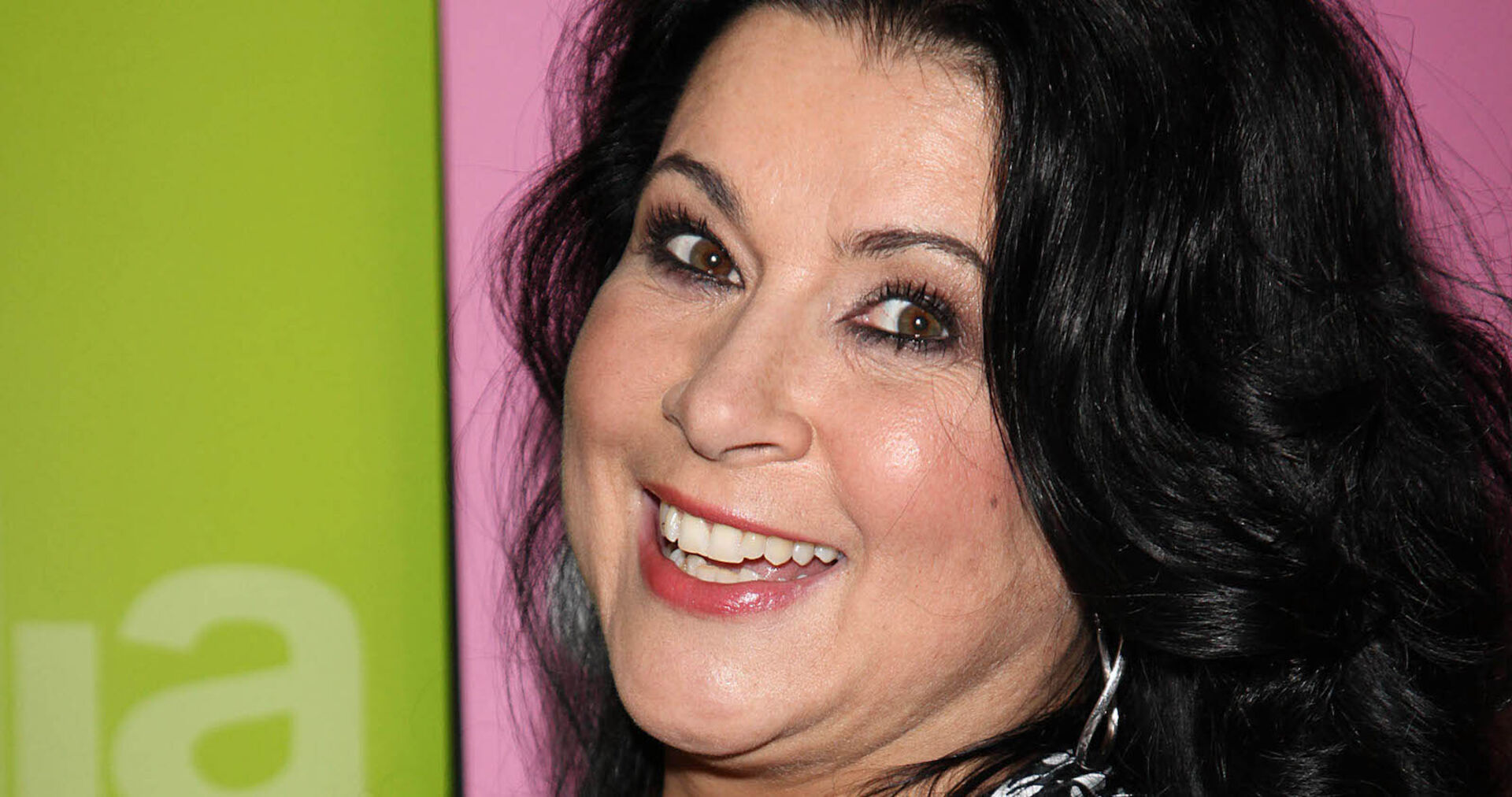 Iris Klein mit offenen schwarzen Haaren vor einer rosa-grünen Leinwand.