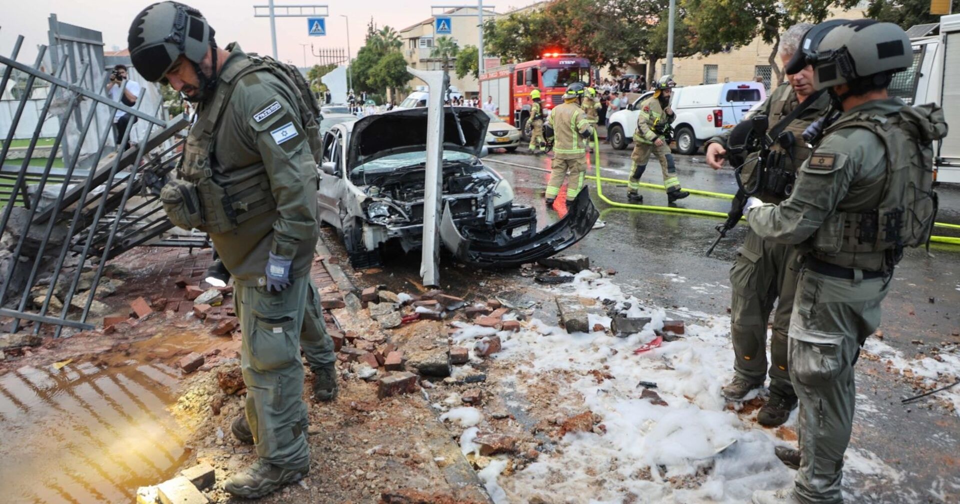 Israelische Polizisten und Rettungskräfte durchsuchen den Ort eines Raketenangriffs. Ein zerbombtes Auto und Trümmer.