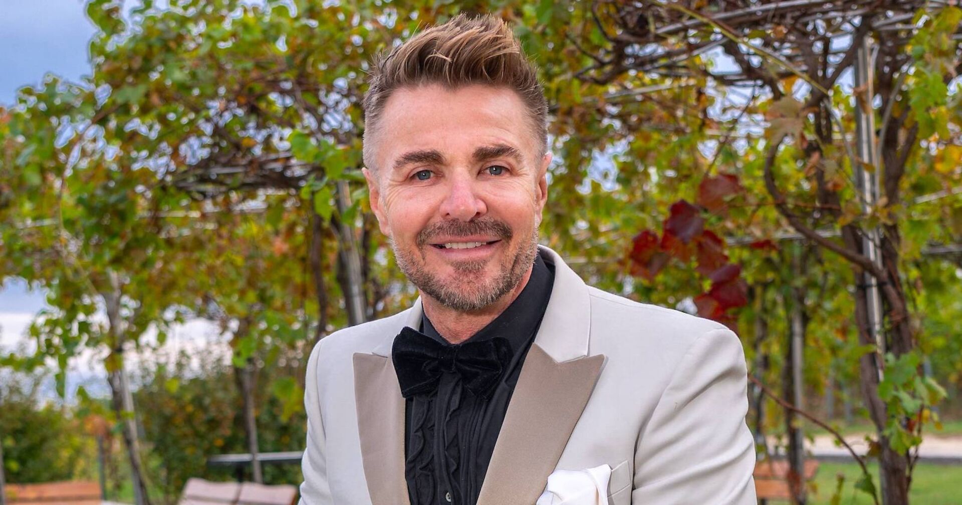 Leo Hillinger mit aufgestellten blonden Haaren und einem grauen Sakko vor einer Weinrebe.