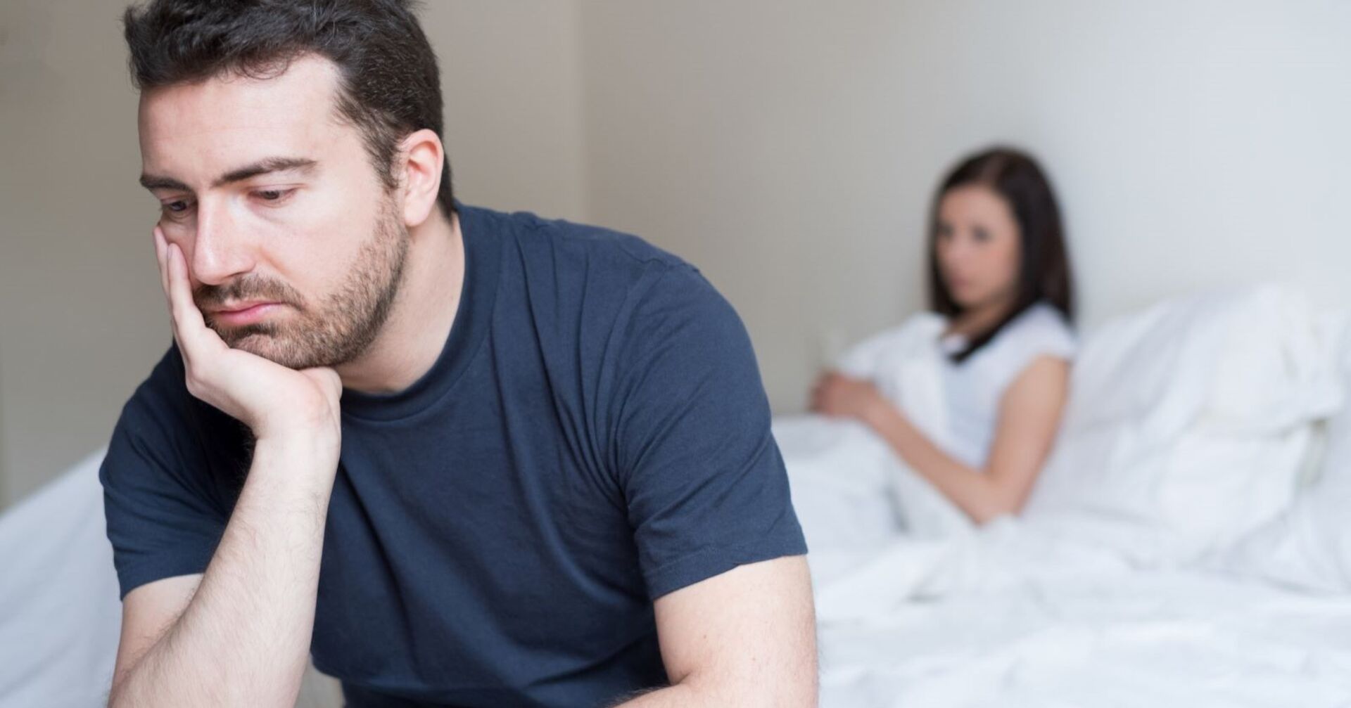 Ein Mann sitzt an den Bettkante, den Kopf in die Hand gestützt. Er wirkt nachdenklich bis bedrückt. Im Bett liegt eine Frau.