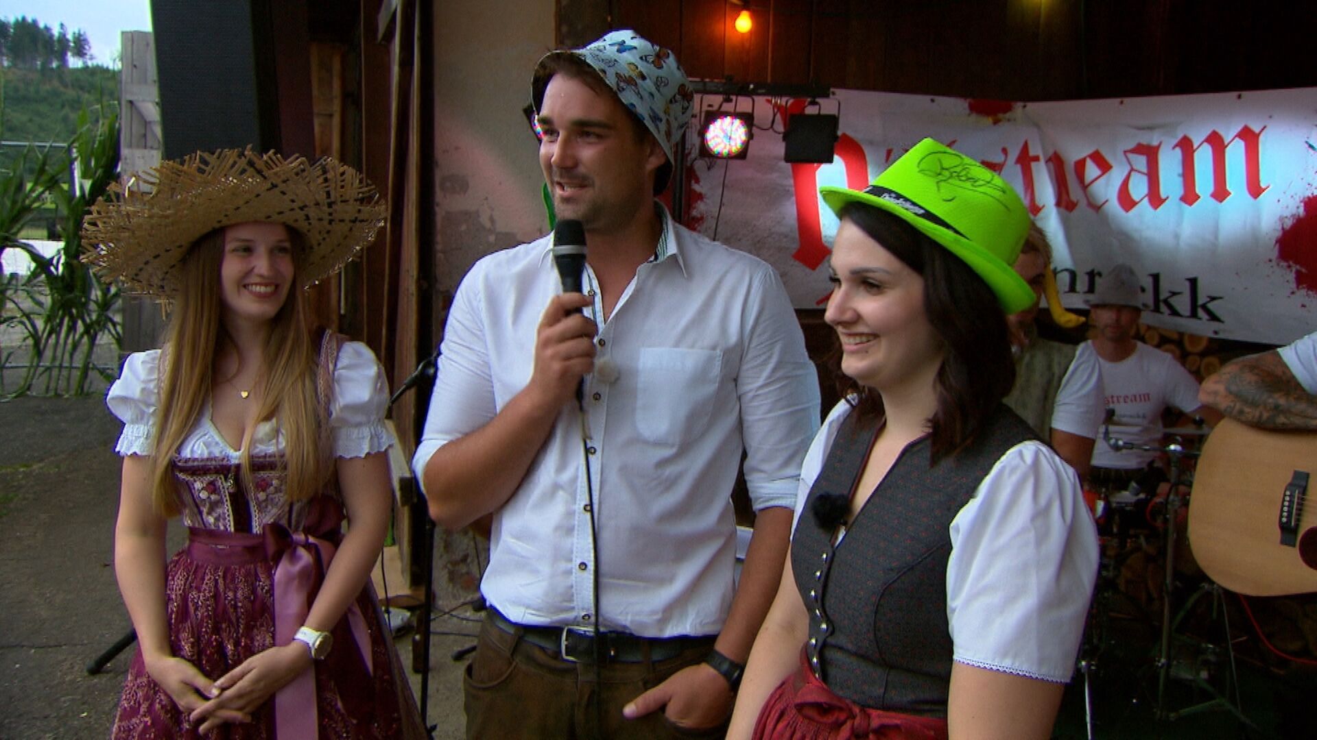 Zwei Frauen stehen neben einem Mann. Sie sind bei einem Fest und er spricht durch ein Mikrofon.