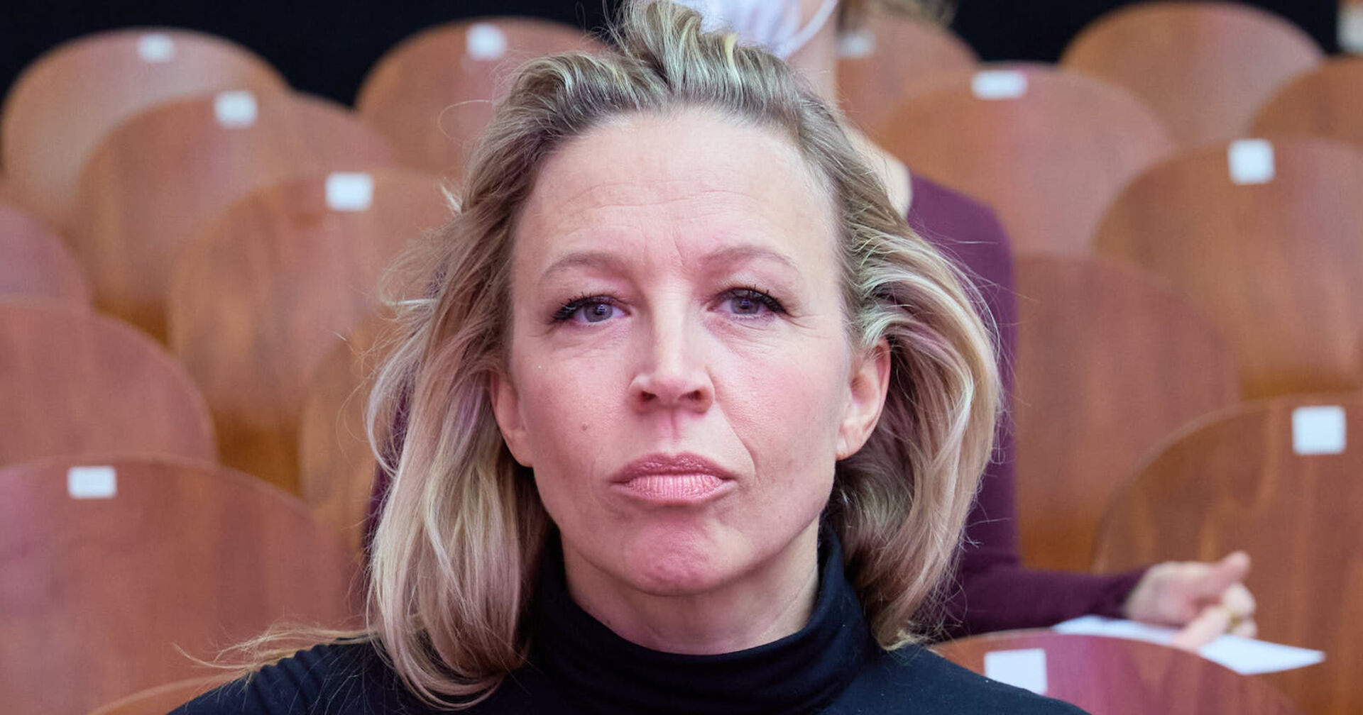 Nina Proll mit offenen blonden Haaren und einem schwarzen Rollkragenpulli sitzt in einer Audience voller Holzstühle.