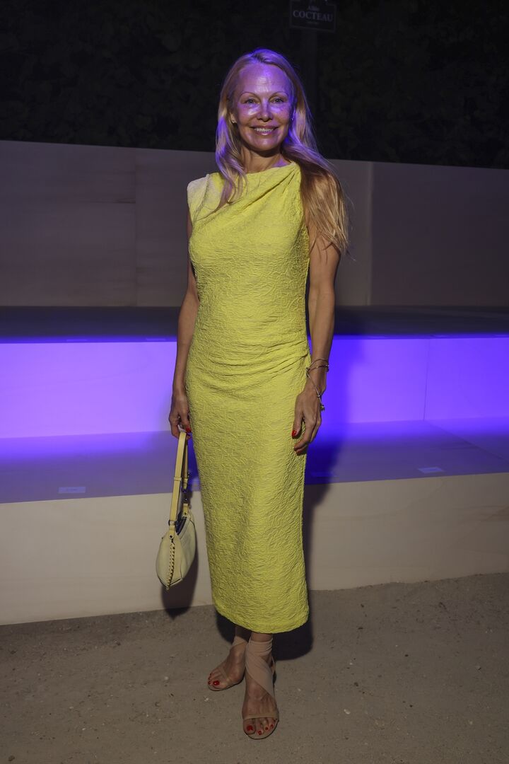 Pamela Anderson mit offenen langen Haare und einem bodenlangen gelebt Kleid. Sie trägt eine gelbe Handtasche.