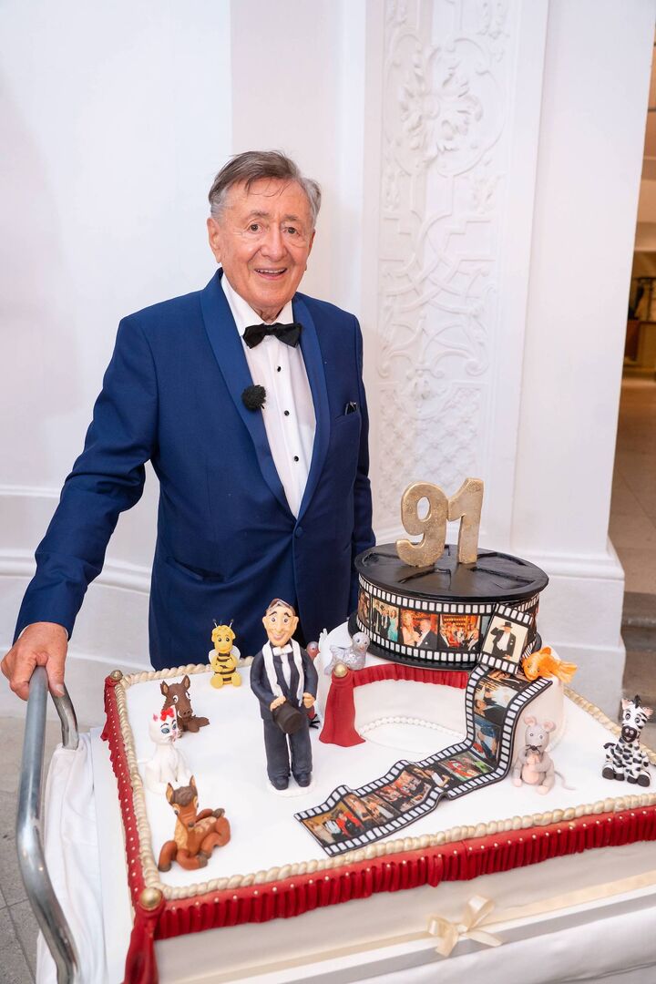 Richard Lugner trägt einen blauen Anzug, eine schwarze Fliege und steht vor einer großen Geburtstagstorte mit viel Verzierung.