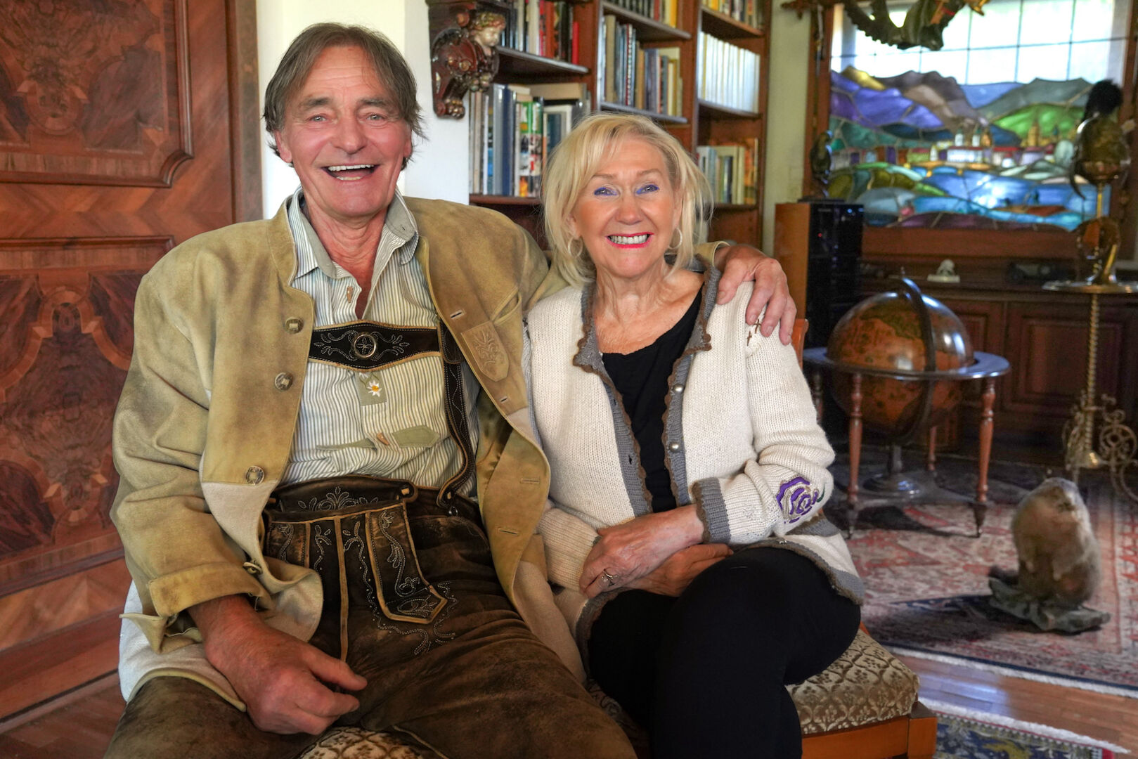 Siggiträgt eine beige Tracht und Rosemarie geschlossene blonde Haare und eine weiße Jacke. Hinter ihnen steht ein ausgestopftes Murmeltier.