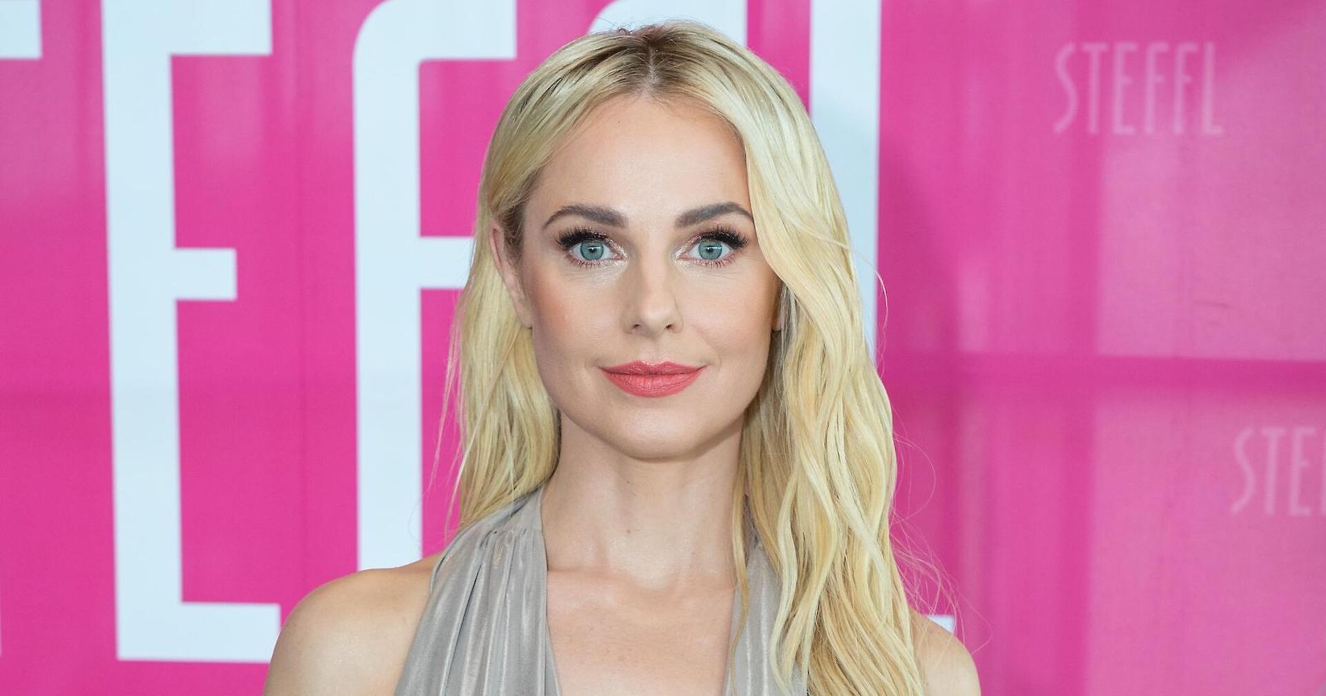 Silvia Schneider mit offenen blonden Haaren vor einer pinken Fotowand.