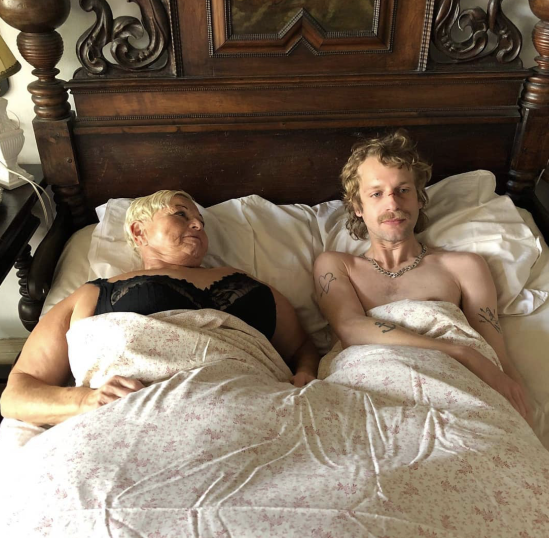 Jazz Gitti, eine reife Damen mit kurzen blonden Haaren und einem schwarzen Oberteil liegt mit dem deutlich jüngeren Voodoo Jürgens (mit Vokuhila) unter der Bettdecke.