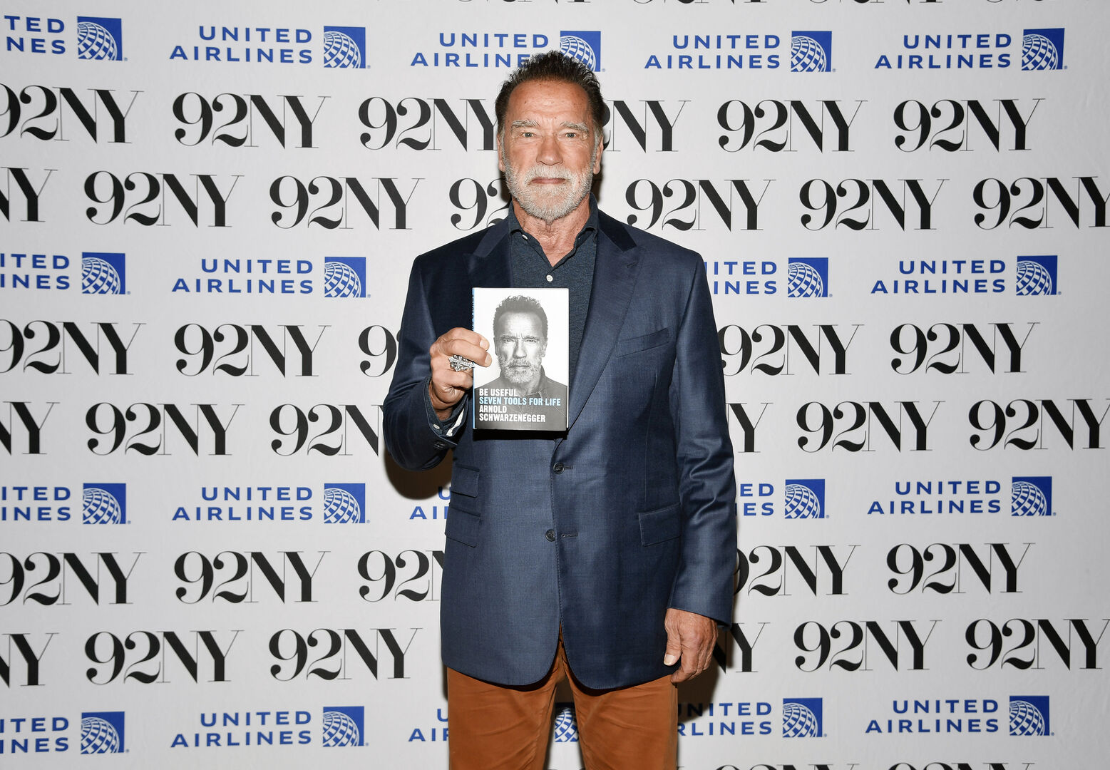 Arnold Schwarzenegger, der auf einem roten Teppich ein Exemplar seines neuen Buches in die Kamera hält