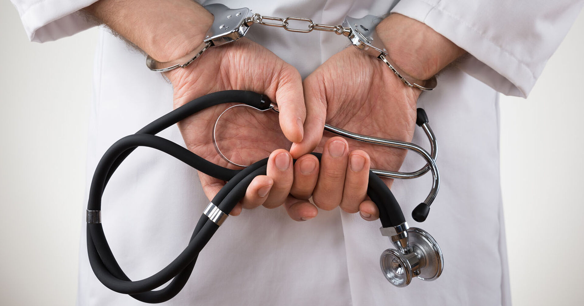Mann im Arztkittel in Handschellen. In den Händen hält er ein Stethoskop.