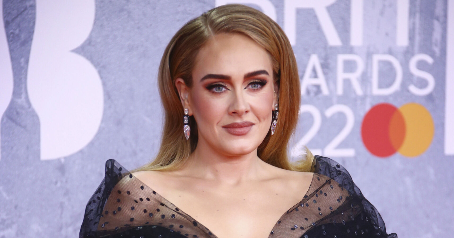 Adele trägt ein schwarzes Kleid mit Spitze und einen tiefen Seitenscheitel. Die Haare sind dunkelblond und offen. Sie steht vor einer violetten Fotowand.