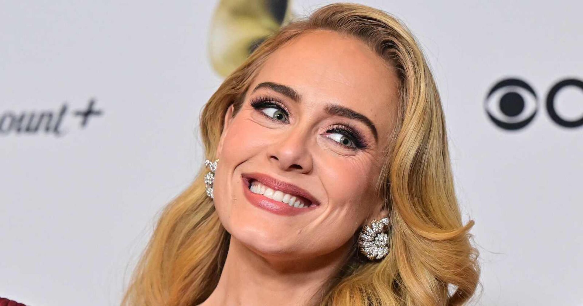 Adele trägt eine voluminöse Föhnfrisur und blonde Haare. Sie ist stark geschminkt im Red-Carpet Look und grinst in die Kamera. Sie trägt auffallende Diamantohrringe.