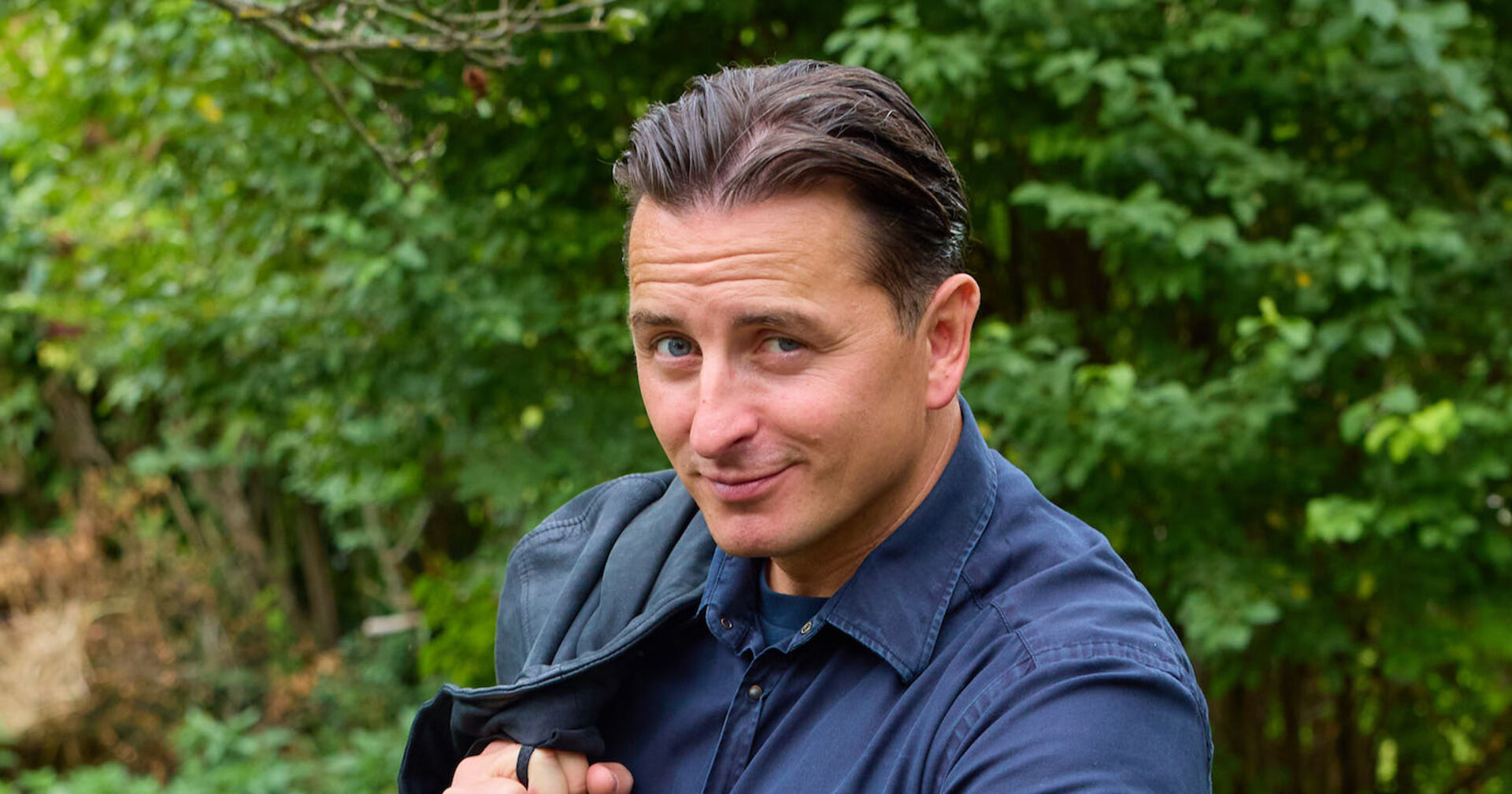 Andreas Gabalier steht vor einem grünen Busch. Er trägt ein dunkelblauen Hemd.