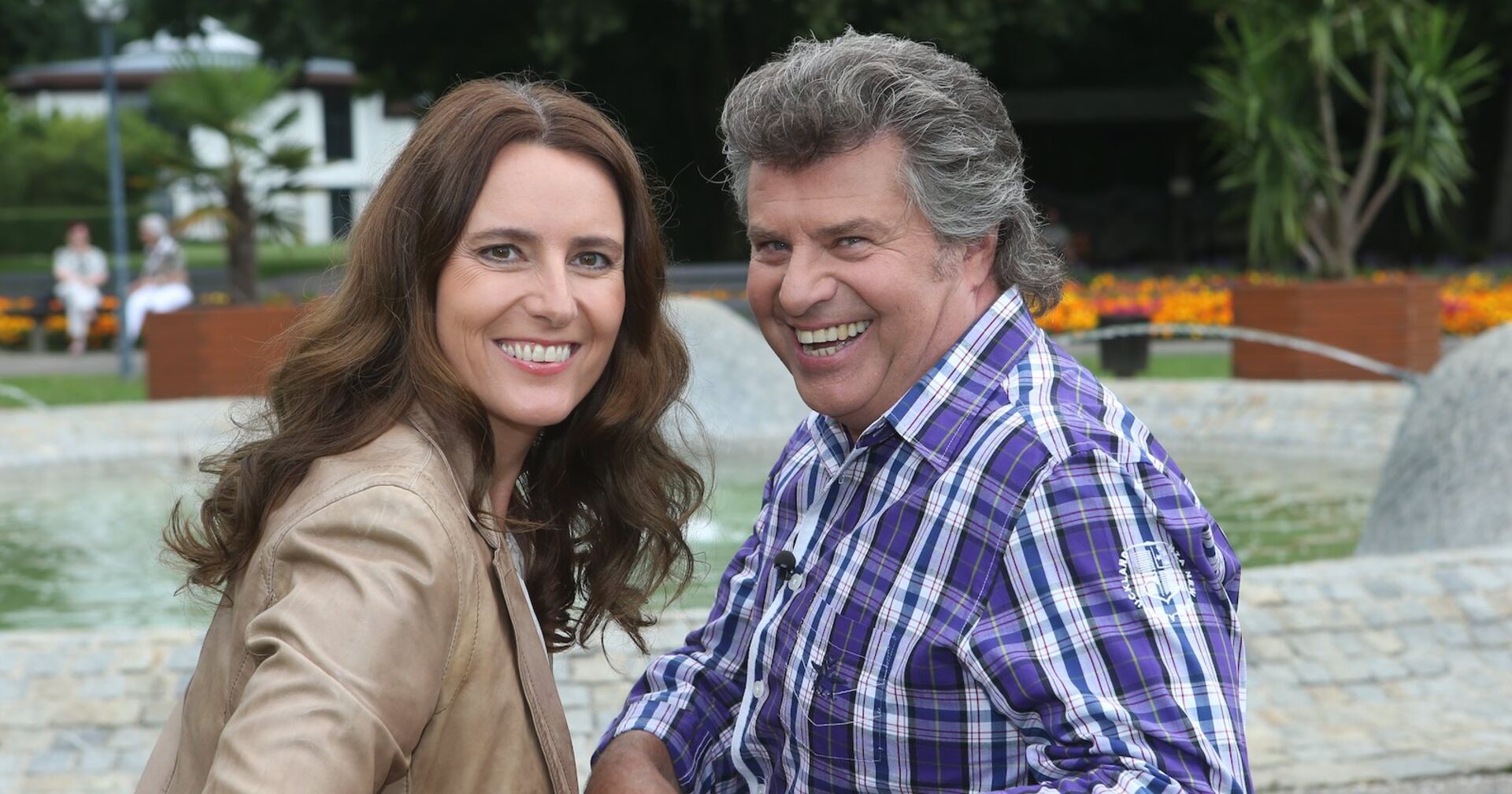 Andy Borg und seine Birgit posen vor einer Wiese auf einer Parkbank. Beide lächeln glücklich in die Kamera.