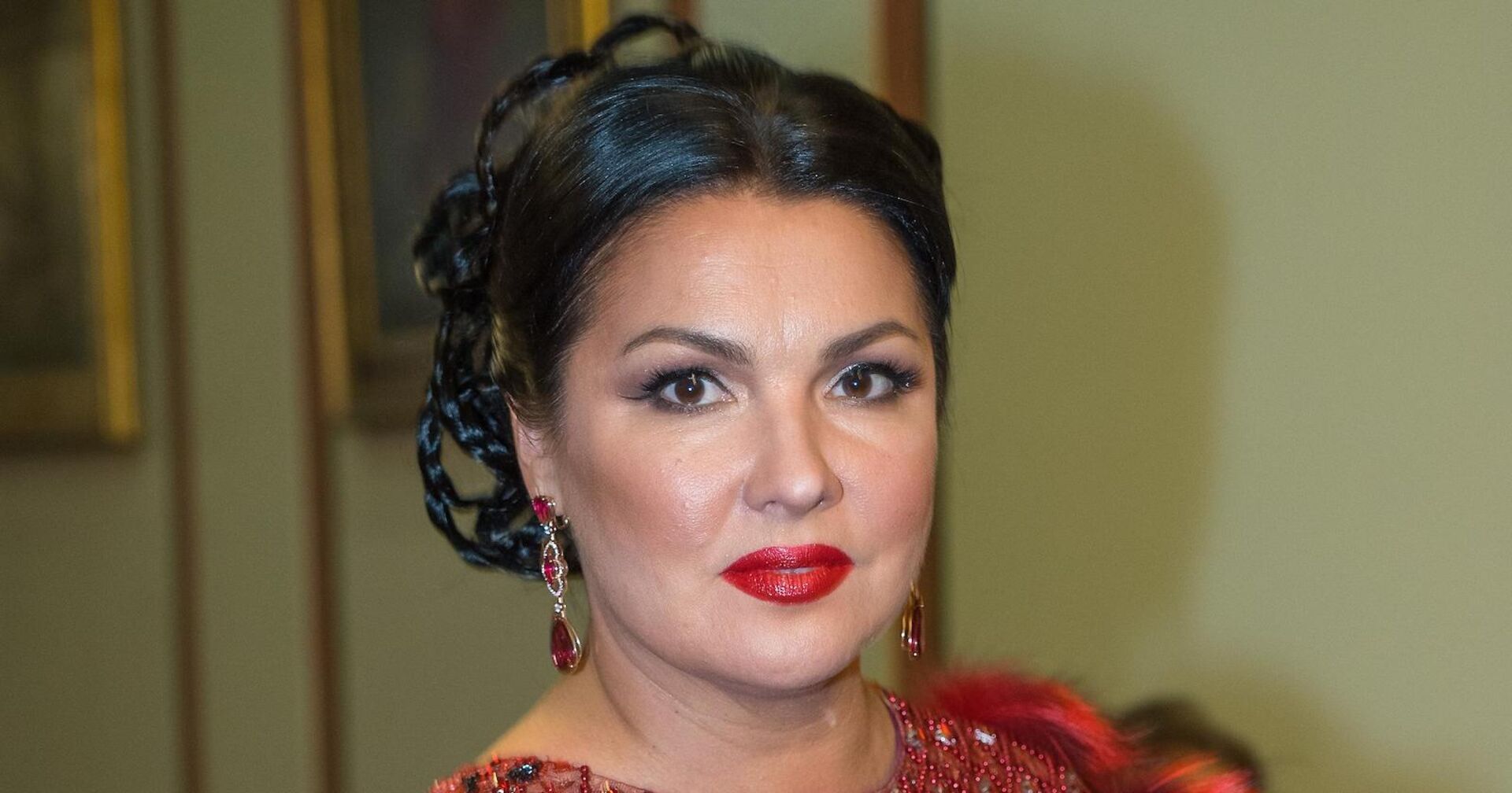 Anna Netrebko trägt hochgesteckte schwarze Haare, stark geschminkte Augen und einen roten Lippenstift.