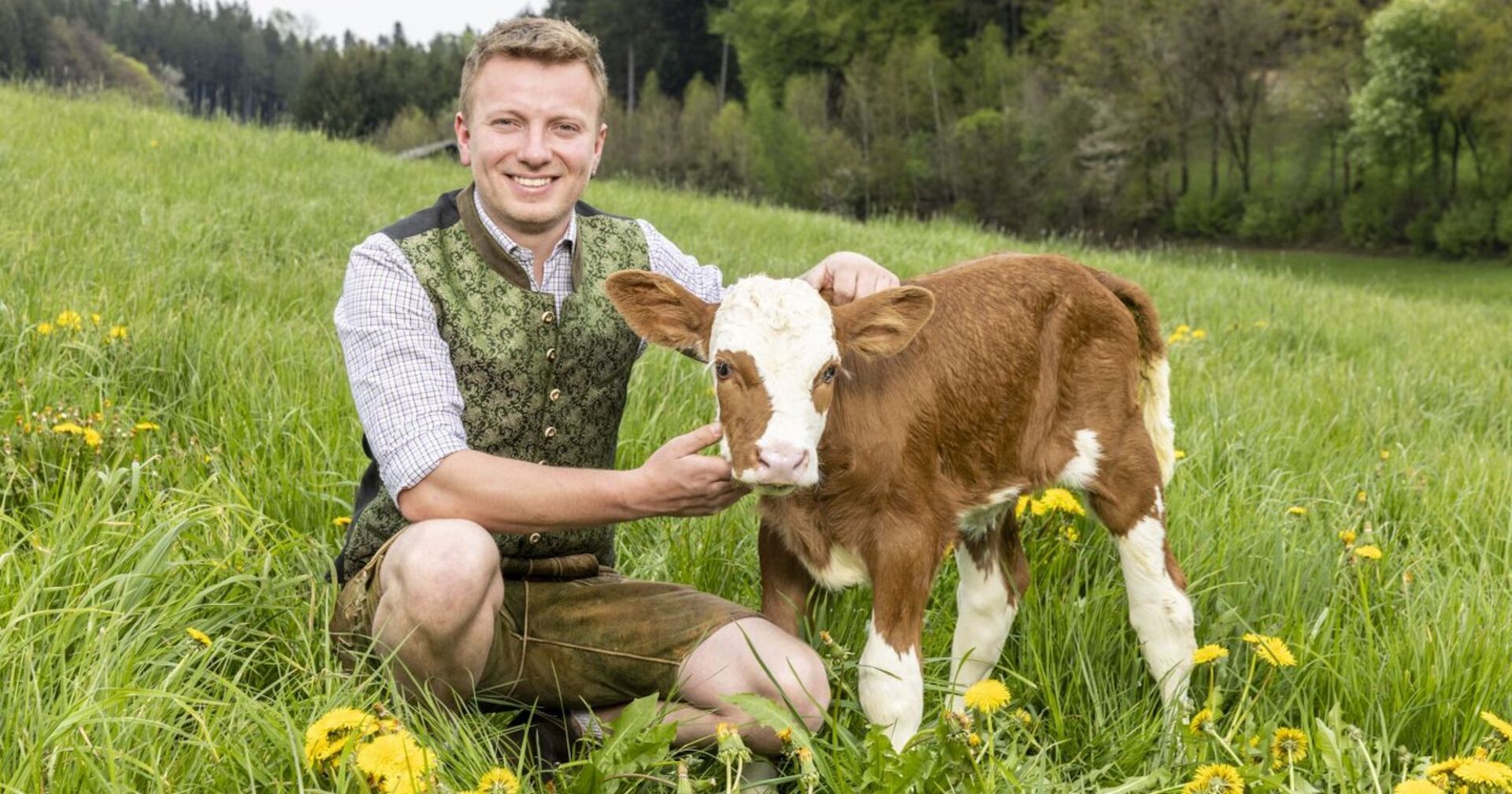 Bauer Johannes kniet auf einer Weide und streichelt ein Kalb. Er trägt eine grüne Tracht und braune Lederhosen.