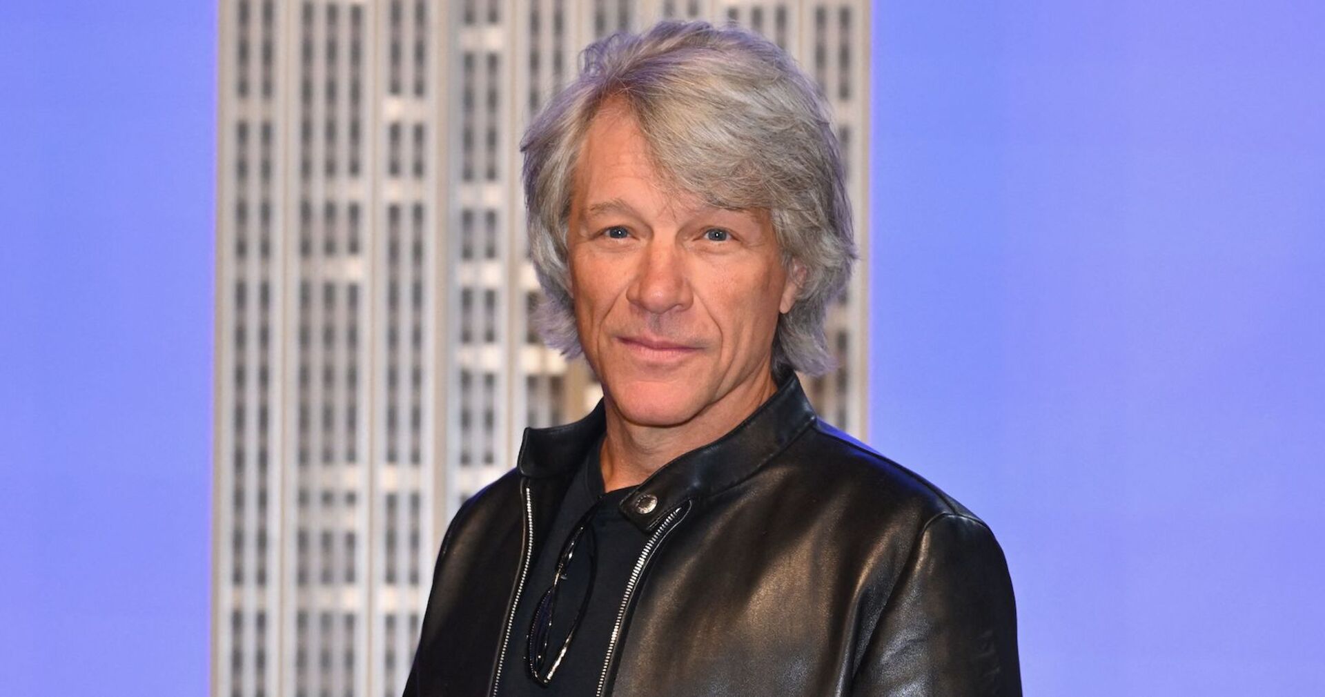 Bon Jovi trägt eine schwarze Lederjacke und steht vor einem hellblauen Hintergrund mit einem Gebäude.