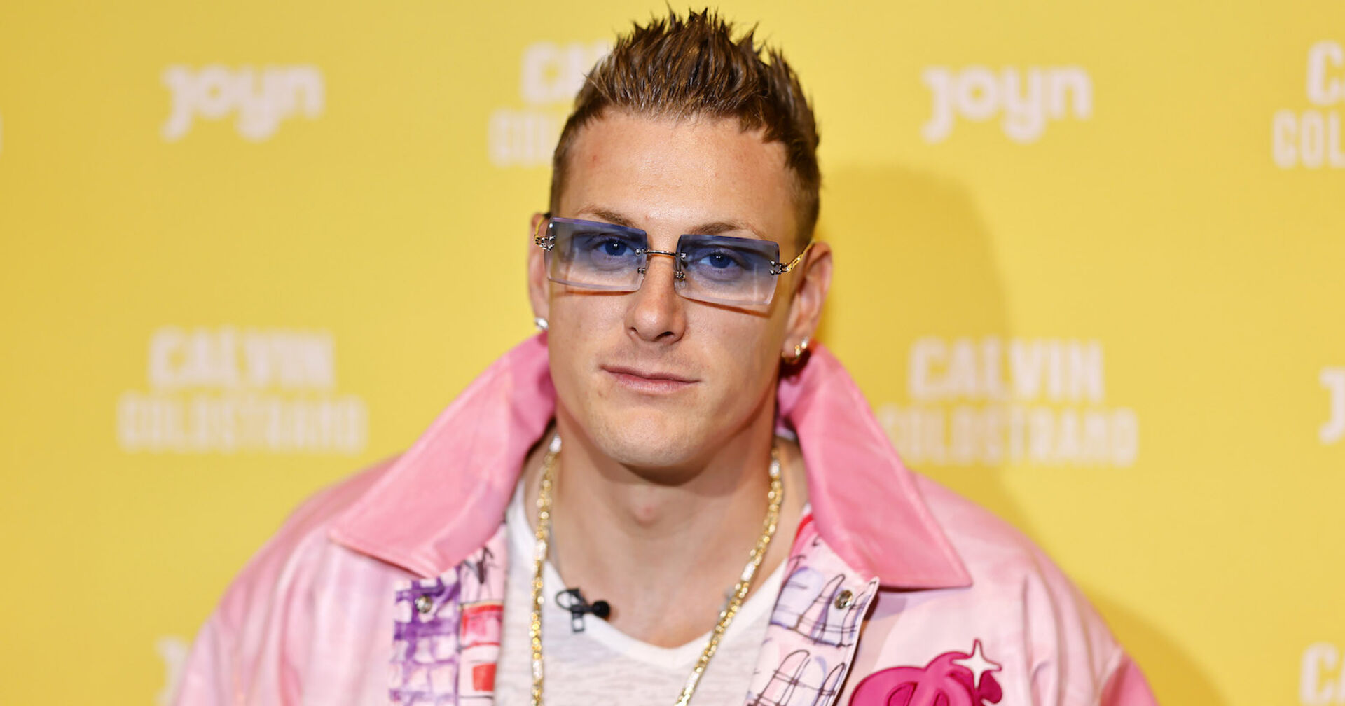 Der Reality Tv Star Calvin Kleinen trägt eine blaue Brille und eine pinke Jacke. Er steht vor einer gelben Fotowand.