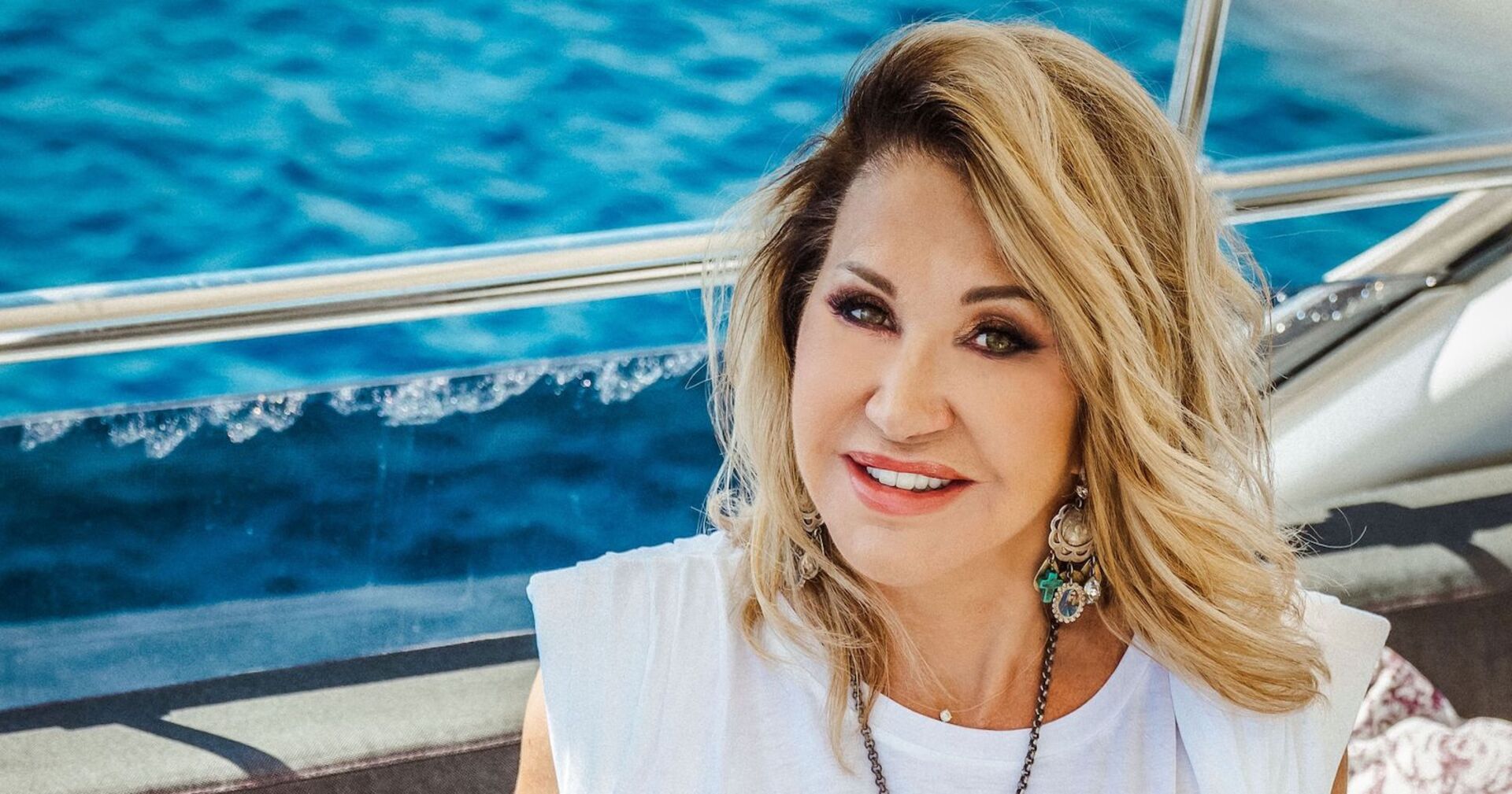 Carmen Geiss hat gewelltes blondes Haar und sitzt auf einer Yacht. Dahinter ist das blaue Meer.