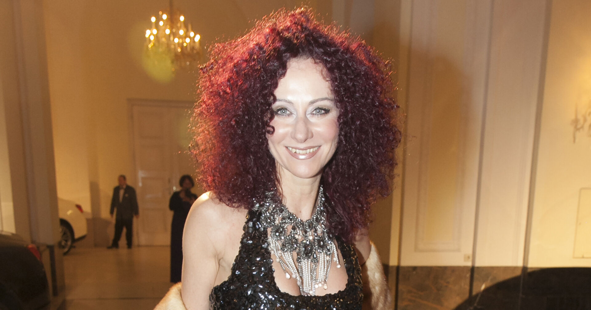 Christine Lugner trägt offene violette Haare und eine prunkvolle Kette. Dazu ein elegantes schwarzes Kleid, das glitzert.