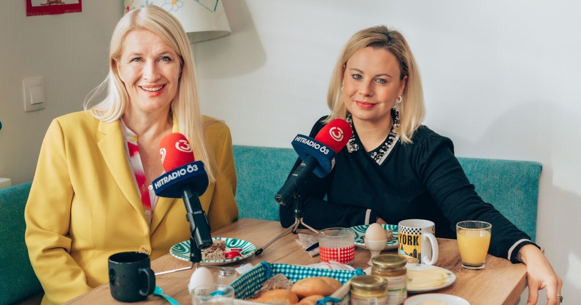 Claudia Stöckl trägt einen gelben Anzug und offene blonde Haare. Katharina Strasser einen dunklen Pullover. Vor ihnen sind zwei rote Mikrofone und ein dicht gedeckter Tisch.