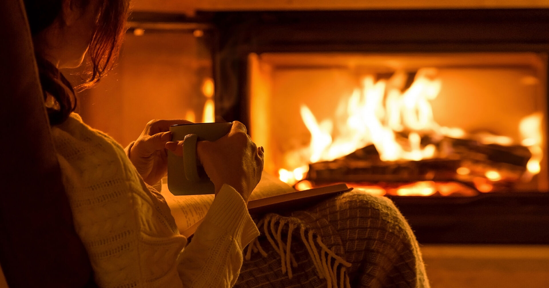 Frau sitzt mit Buch vor einem Kamin | Credit: iStock.com/Rasulovs