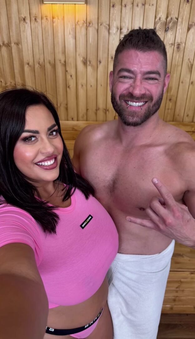 Costina trägt ein rosa Top und lacht in die Kamera. Neben ihr steht ein Pornodarsteller aus Amerika mit einem weißen Handtuch um die Hüfte.