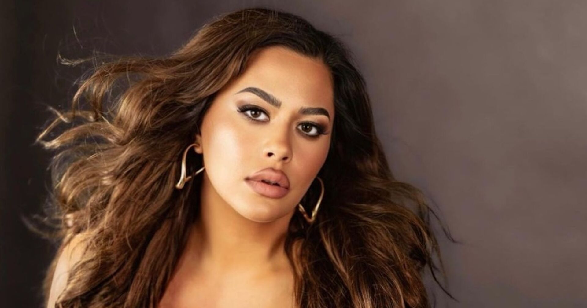 Costina trägt offene, gewellte Haare und ein starkes Make Up, sowie goldene große Ohrringe. Sie posiert vor der Kamera. Hinter ihr ist eine dunkelgraue Fotowand.