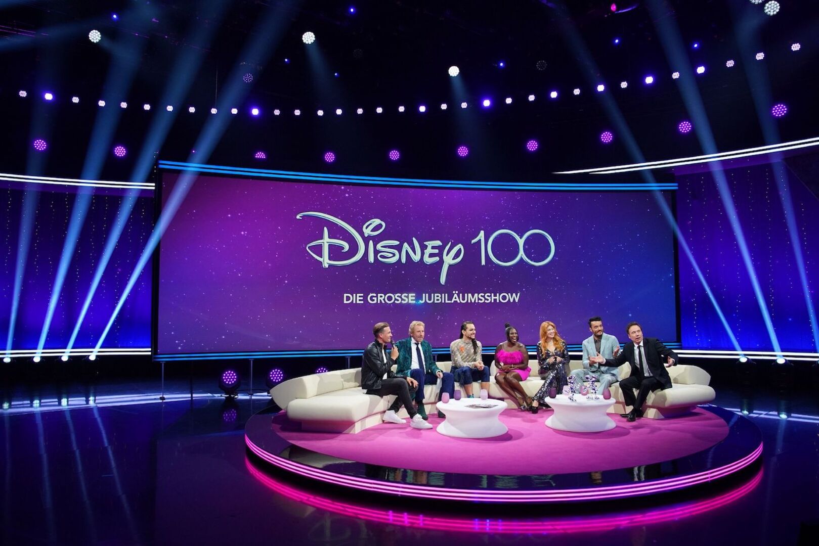 Prominente Gäste auf einer beigen Couch. Dahinter eine Leinwand auf der steht&quot; Disney 100&quot;.