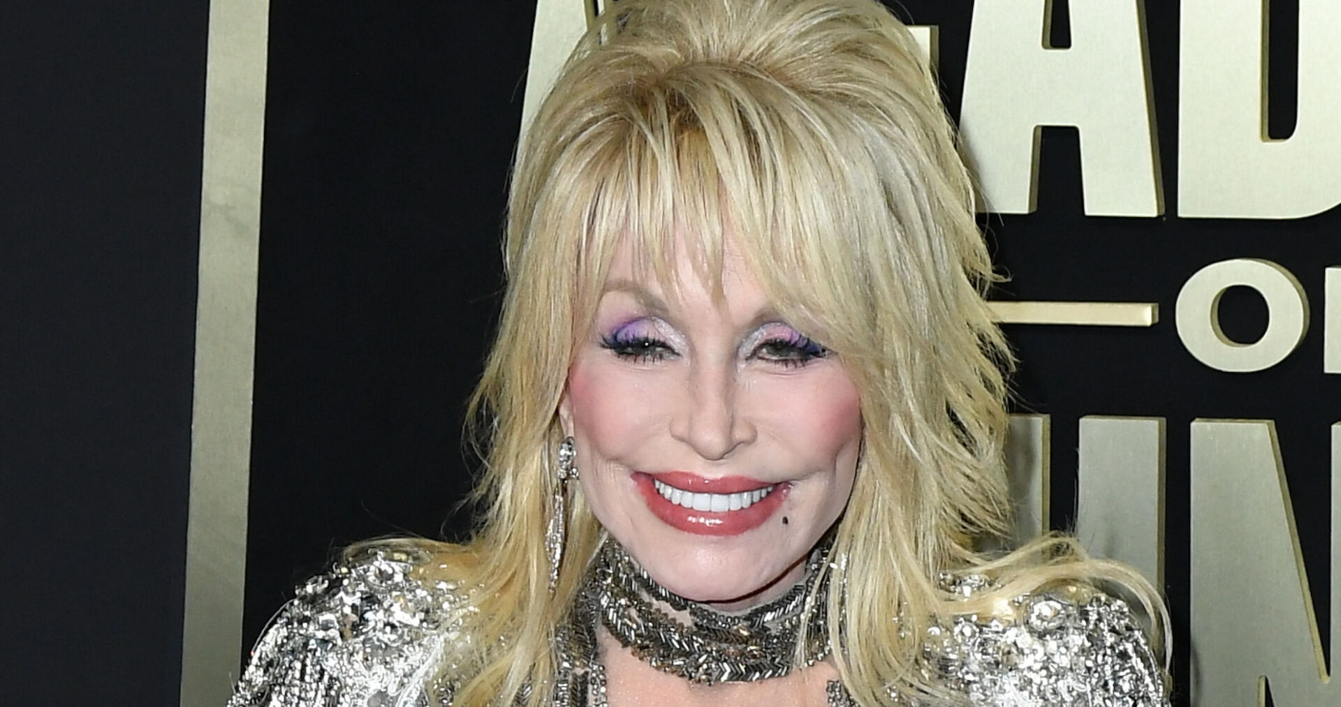 Dolly Parton hat blonde, toupierte Haare, viel Make-up und ein glitzerndes Kleid. Sie steht vor einer schwarzen Fotowand.