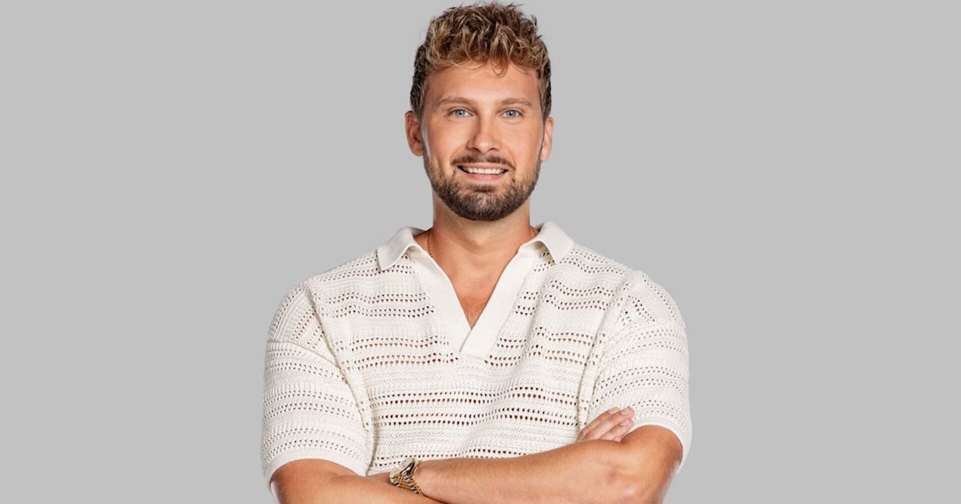 Ex-Bachelor Dominik Stuckmann trägt ein weißes T-Shirt und verschränkt die Hände. Er steht vor einer grauen Wand.