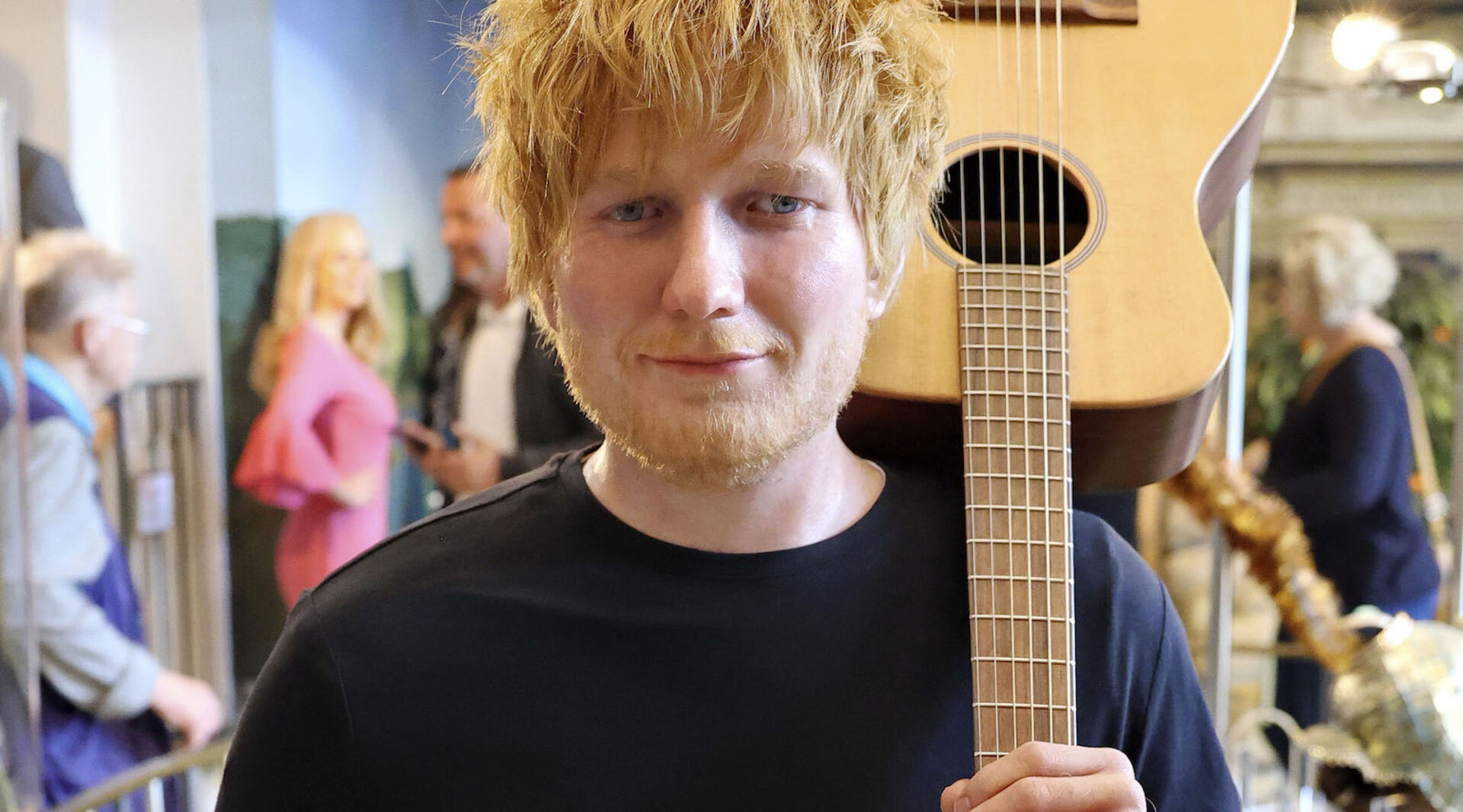Ed Sheeran trägt ein schwarzes T-Shirt und hält eine Gitarre auf den Schultern.