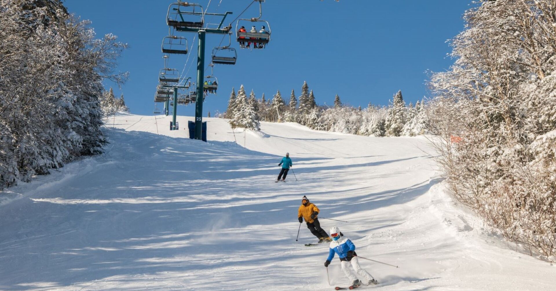Drei Skifahrer auf einer Piste, blauer Himmel, darüber ein Sessellift.
