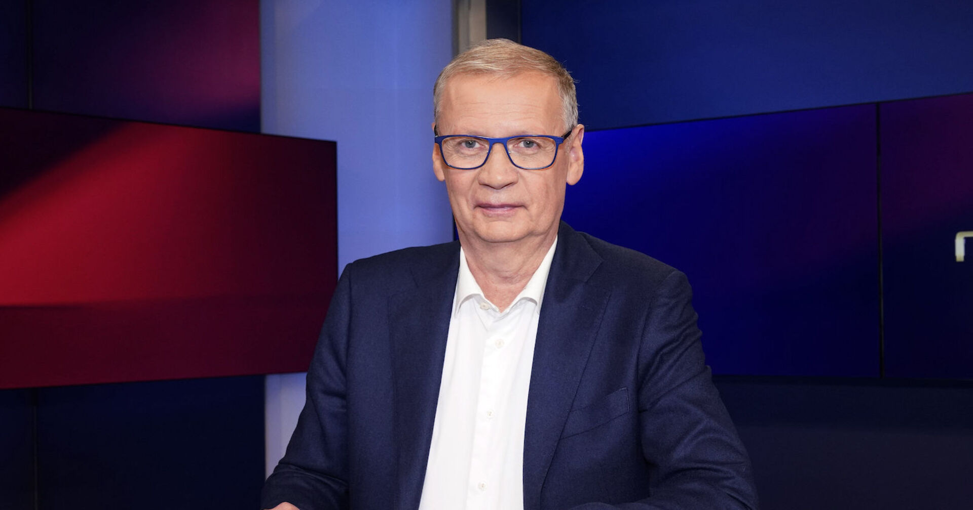 Günther Jauch trägt einen dunkelblauen Anzug und eine weiße Bluse. Er sitzt vor einem blau-roten Hintergrund.