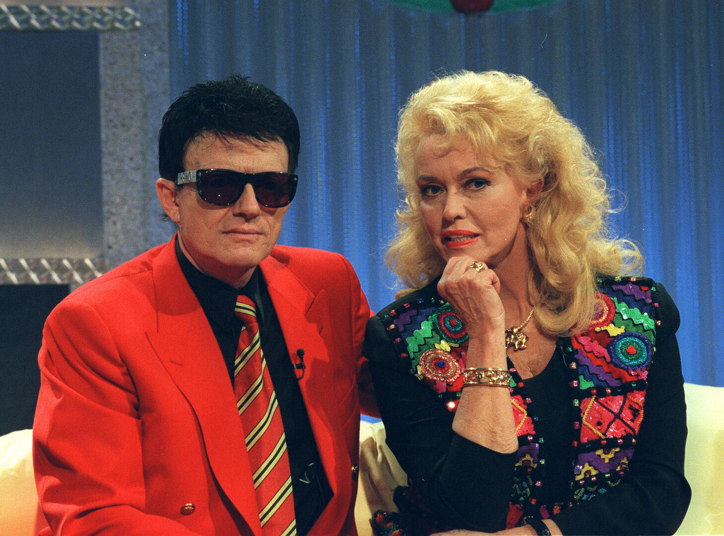 Heino trägt schwarze Haare und einen roten Anzug und eine Sonnenbrille. Hannelore welliges blondes Haar und ein buntes Oberteil.