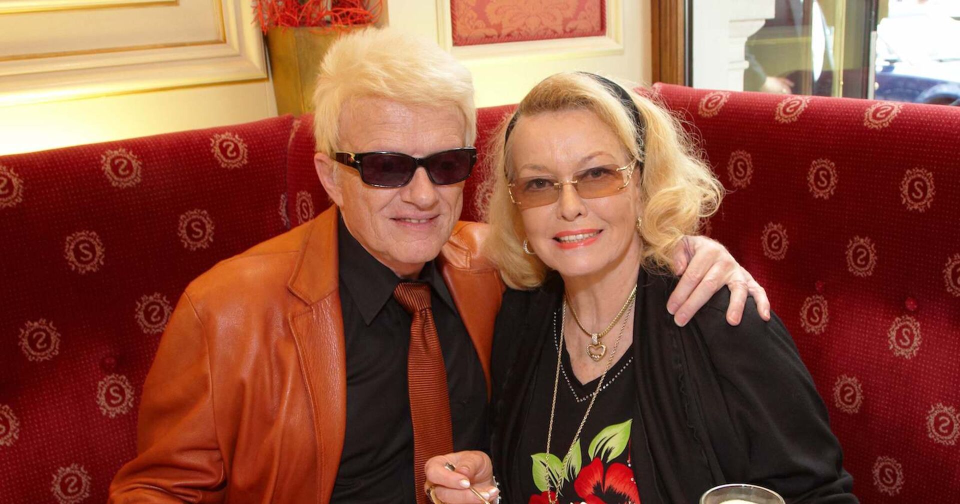 Heino trägt eine orangene Jacke und ein schwarzes Hemd, sowie eine orangene Krawatte. Hannelore eine schwarze Blue. Heino trägt auch eine Sonnenbrille. Beide sitzen gemeinsam am Tisch.