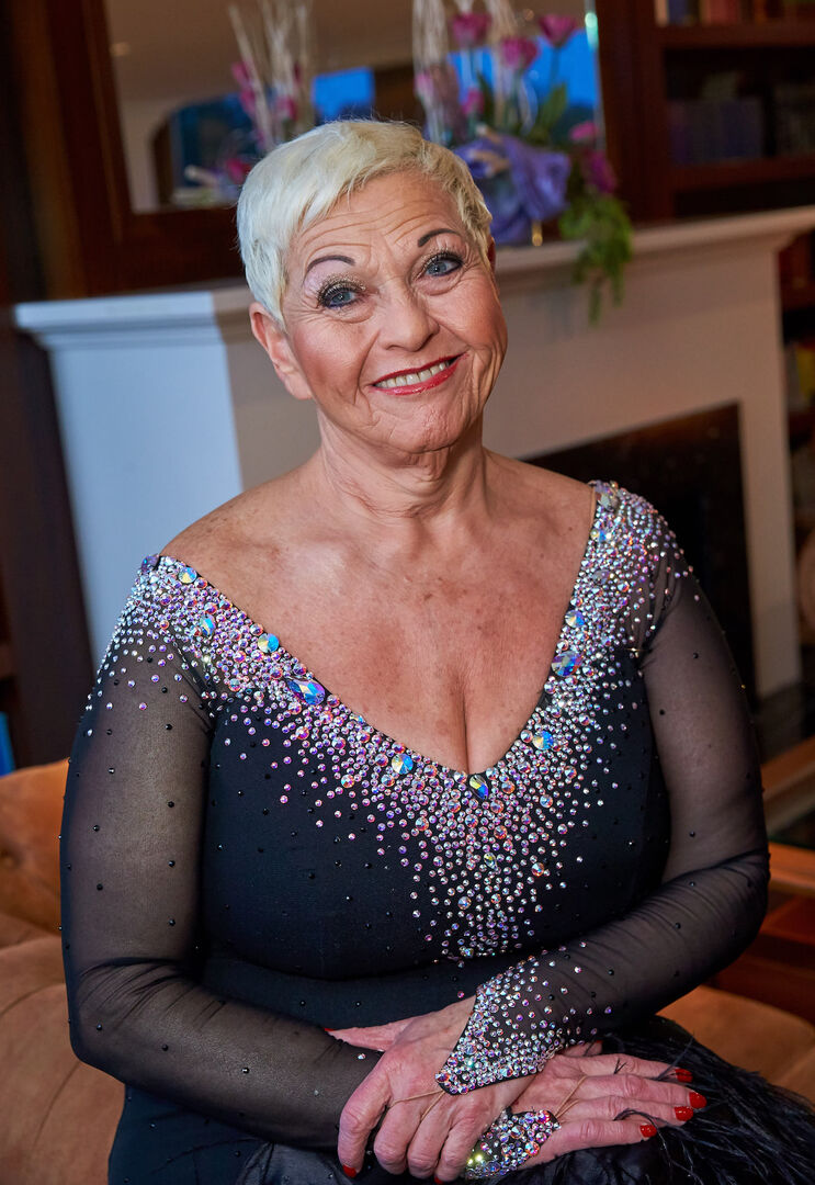 Jazz Gitti in einem schwarzen Ballkleid mit kurzen blonden Haaren. Sie lächelt in die Kamera und sitzt auf einem Stuhl. Das Dekoltee ist mit Glitzersteinen verziert.