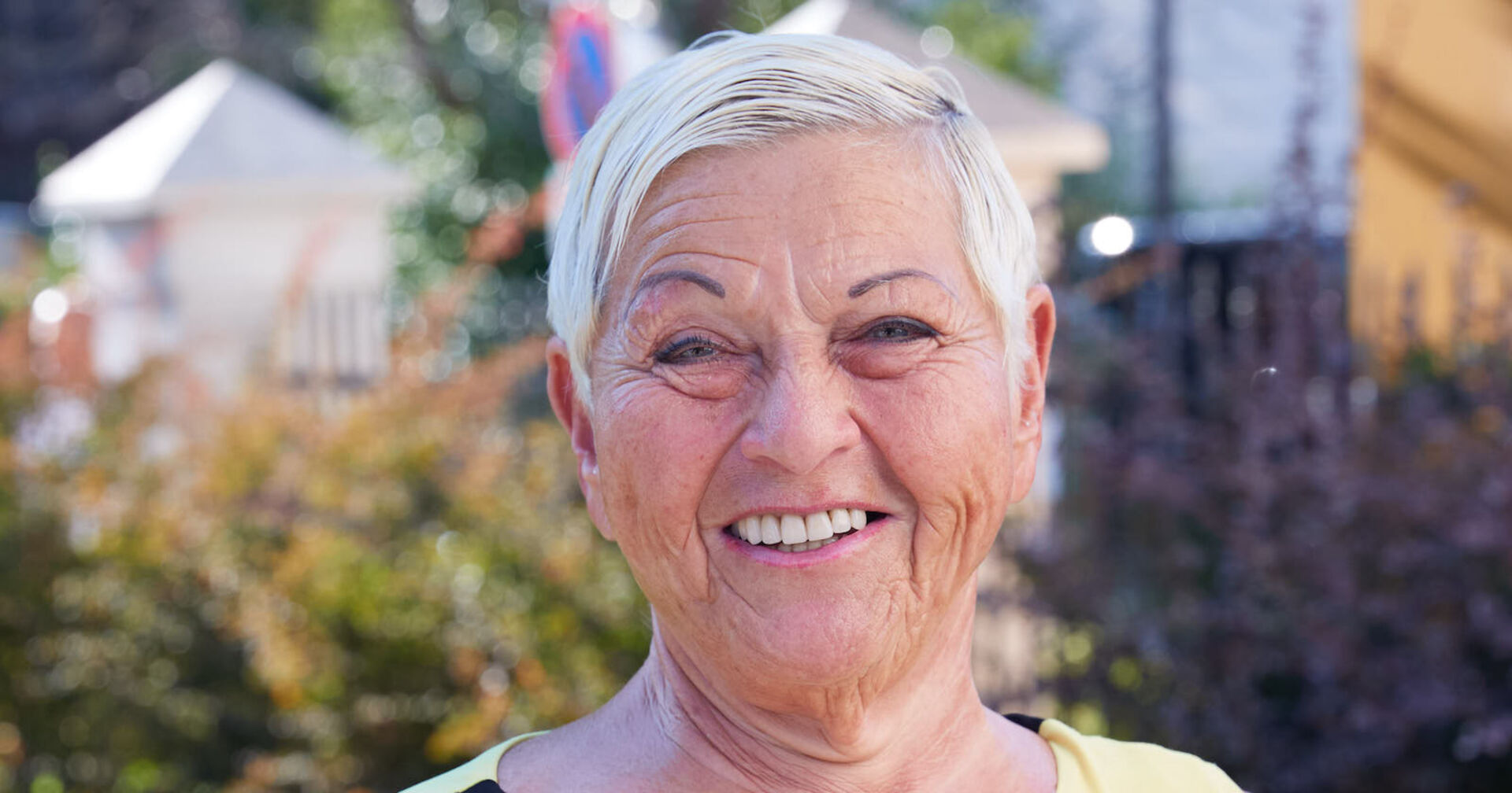 Jazz Gitti steht vor einer Wiese und Häusern. Sie trägt kurze platinblonde Haare und lächelt in die Kamera.