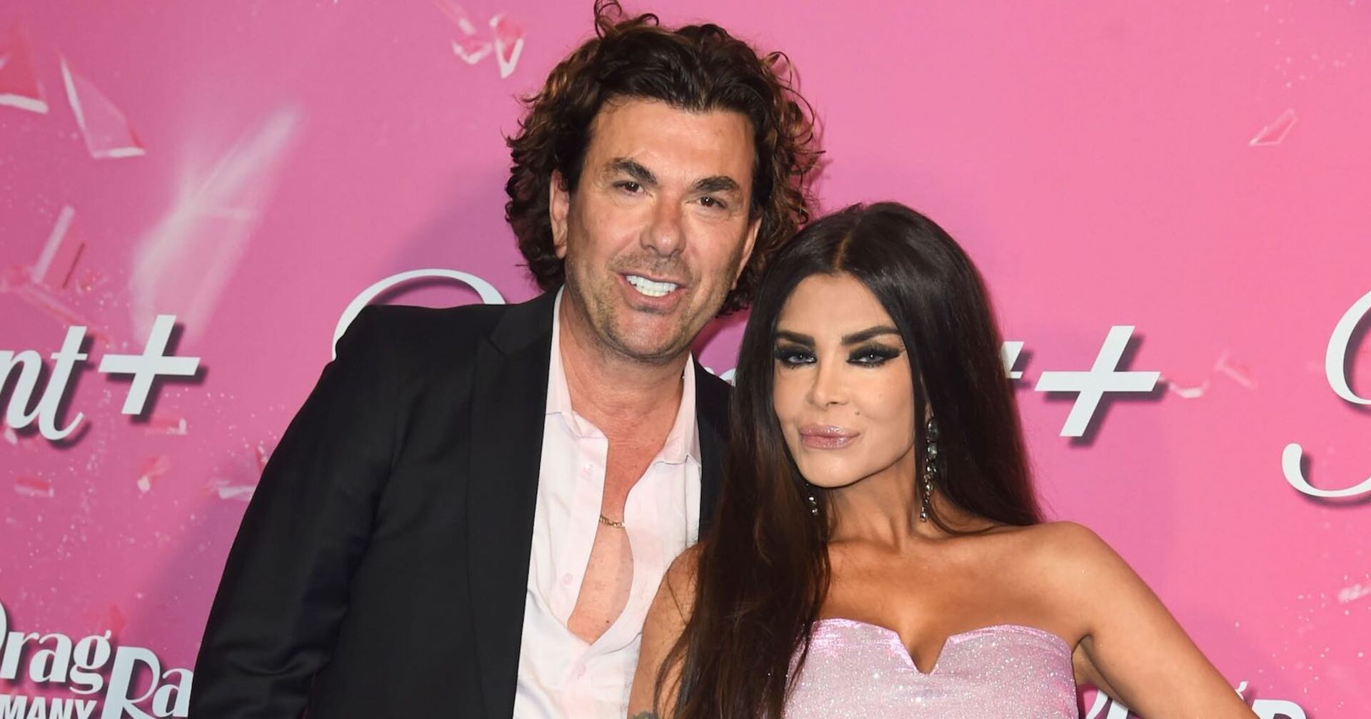 Kader Loth trägt ein rosa Kleid ohne Träger und lehnt an ihrem Mann Isi, er trägt einen schwarzen Blazer. Beide stehen vor einer Fotoleinwand.